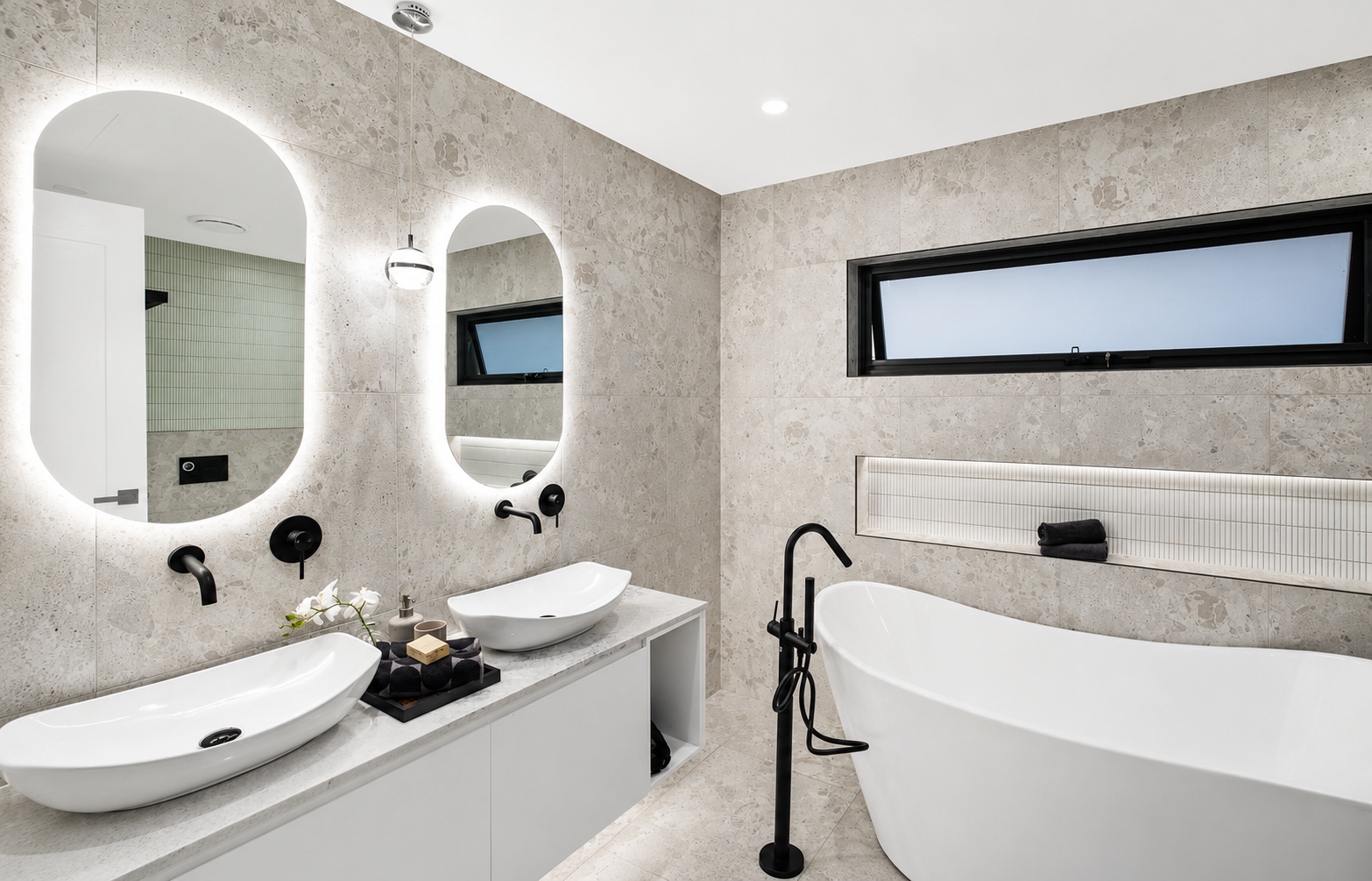 Modern bathroom with two vessel sinks, backlit oval mirrors, a black freestanding tub, and a horizontal window.