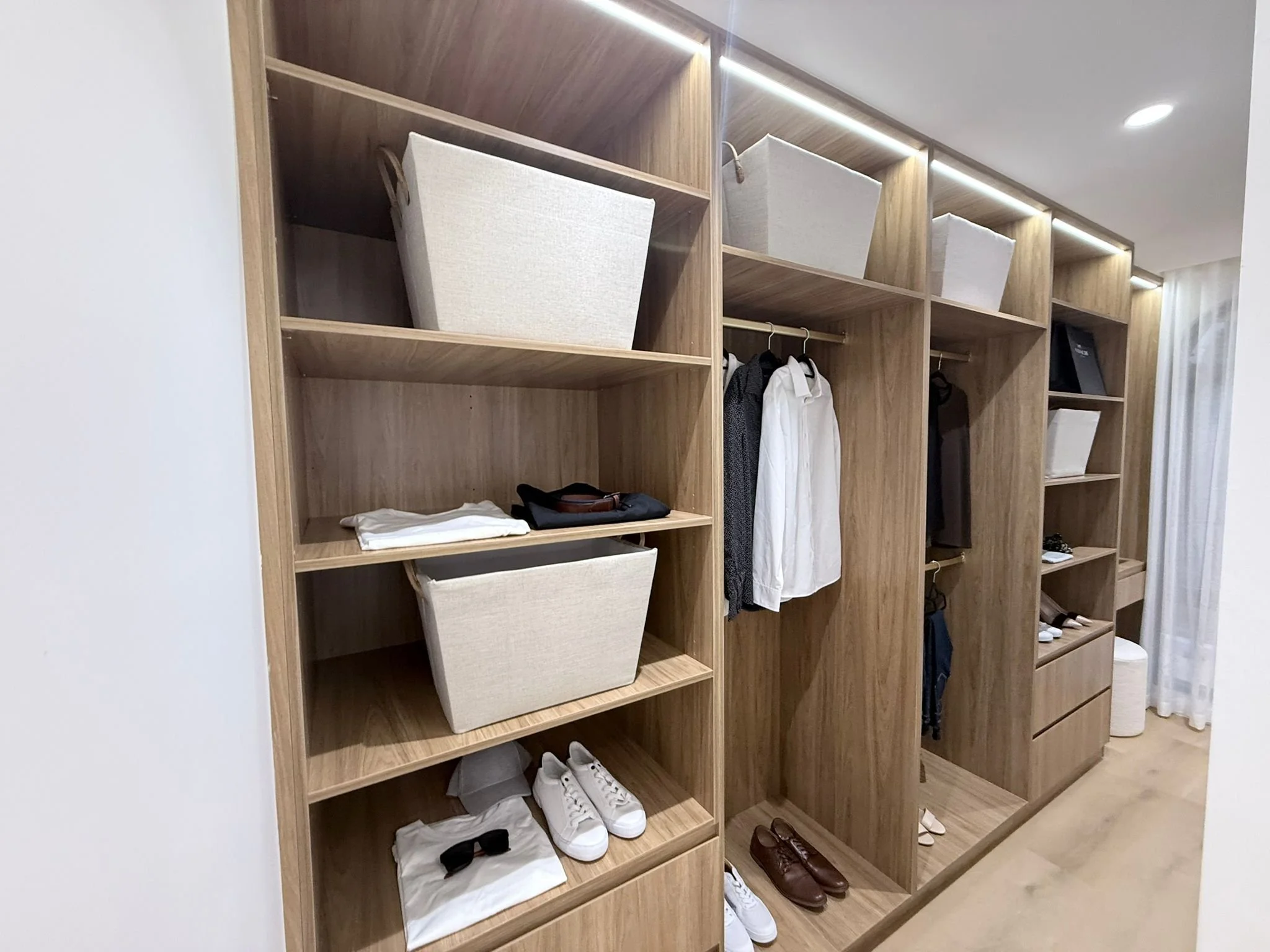 Wooden closet with open shelves and hanging space, containing shoes, folded clothes, bags, and other personal items.
