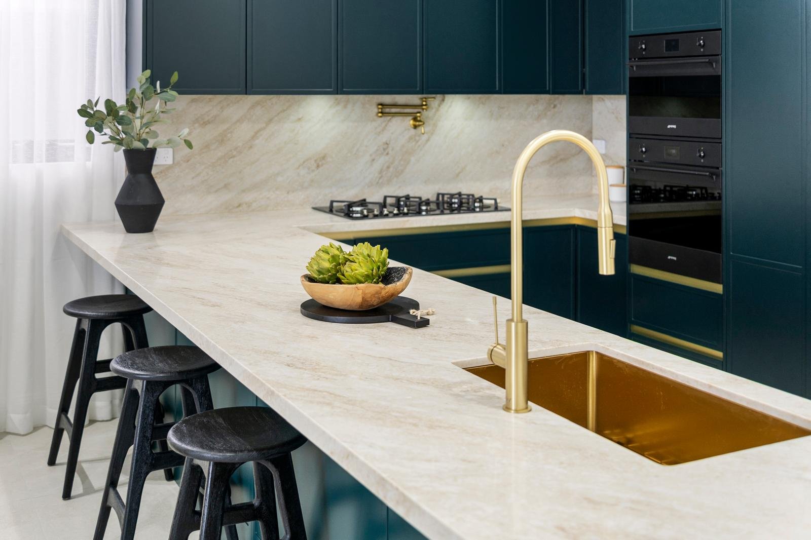 modern kitchen with blue cabinets, marble countertop, gold faucet, black bar stools, decorative bowl with green artichokes, potted plant, built-in stove, and double ovens.