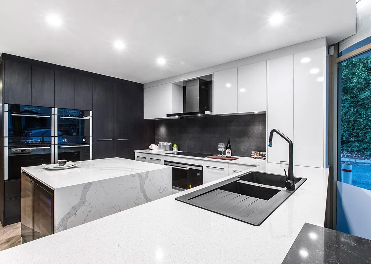 Modern kitchen with black and white cabinetry, marble island, black sink, and built-in ovens.