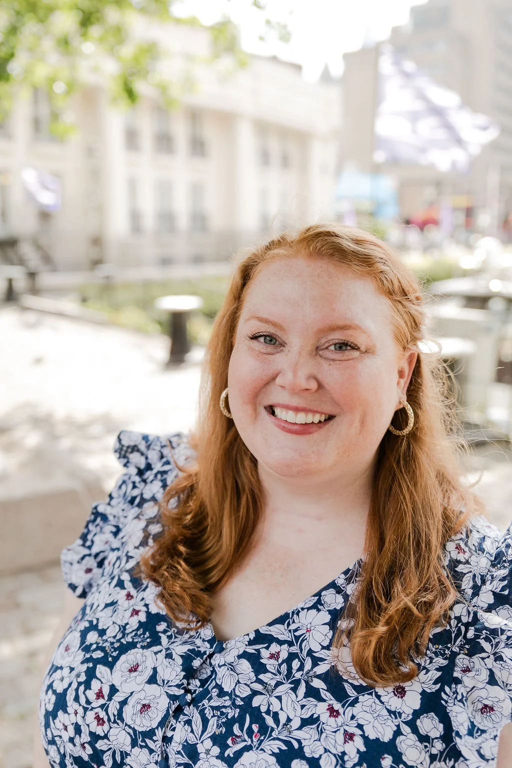 A woman with red hair smiling outdoors in front of blurred buildings and trees.