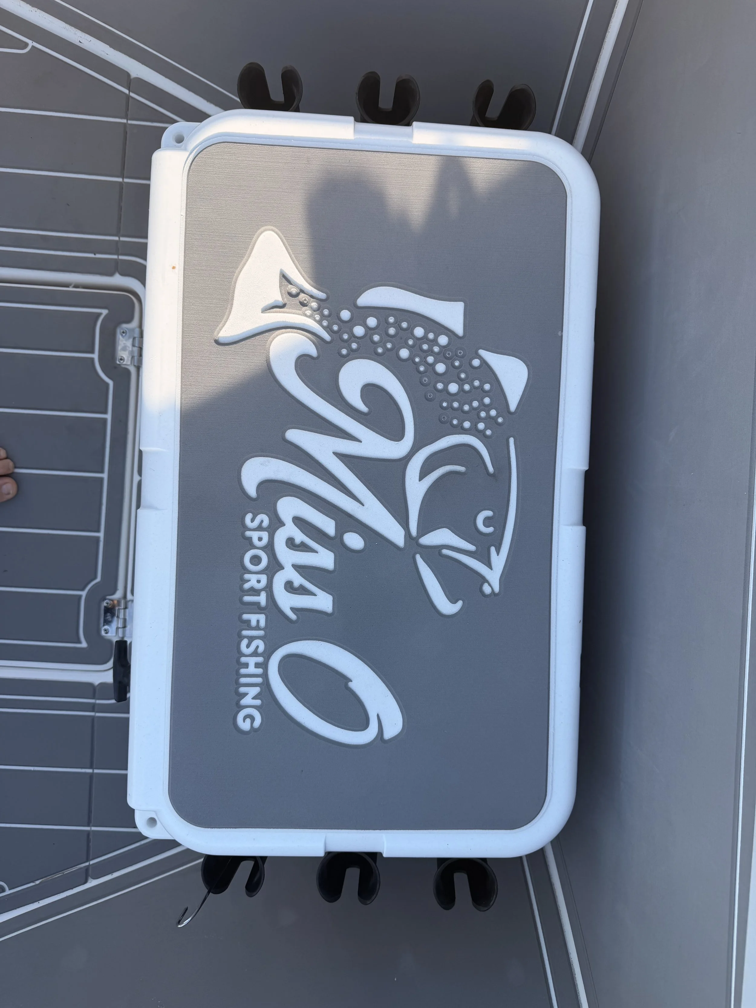 A rectangular fishing tackle box with a logo of a fish and the words 'Sport Fishing' on the lid, resting on a surface with rails and hooks underneath.