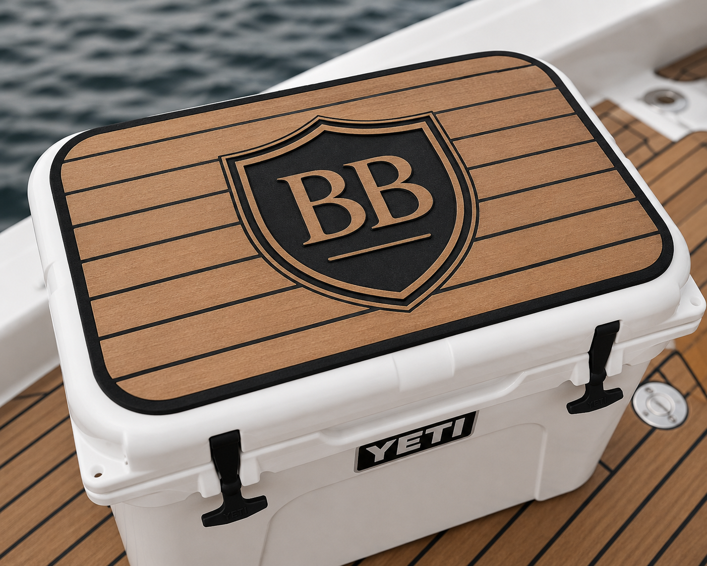 A white YETI cooler with a wooden lid that has a black shield emblem and the letters 'BB' on top. The cooler is on a boat with wooden decking, and water can be seen in the background.