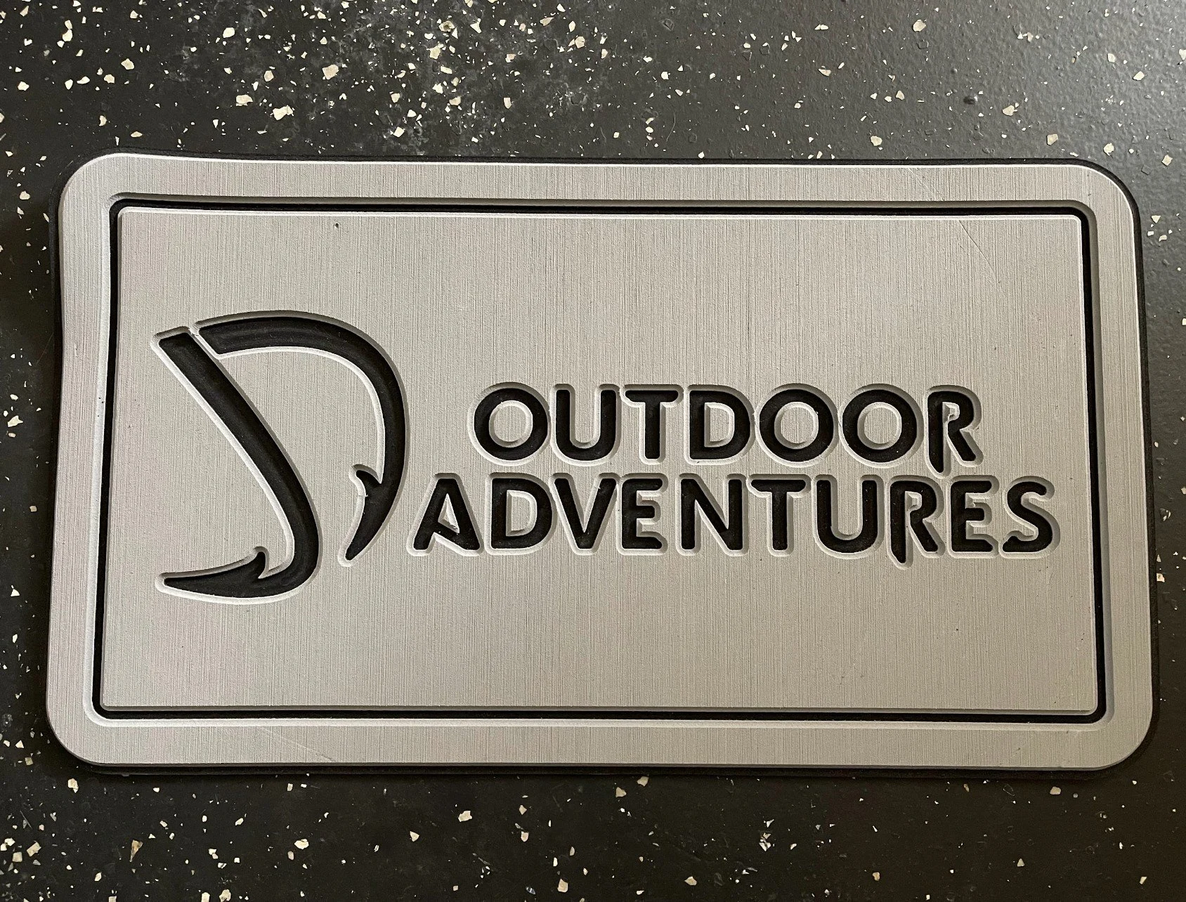 Metal sign with a fish logo and the words "OUTDOOR ADVENTURES" engraved in black, set on a black speckled surface.