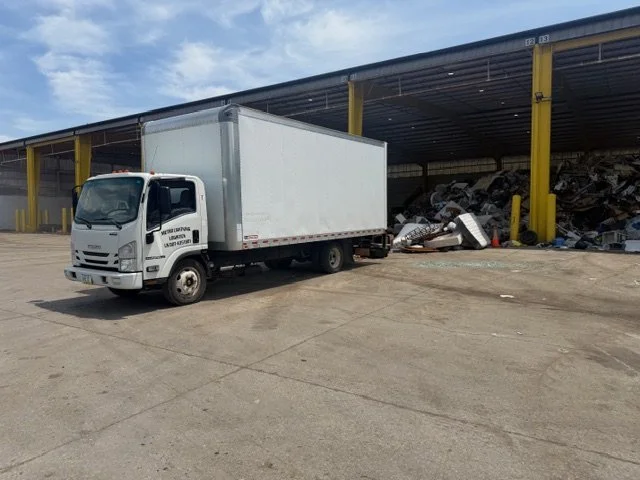 A white box truck parked on a concrete lot near a large scrap yard with piled metal and vehicles under a covered warehouse with yellow support beams.