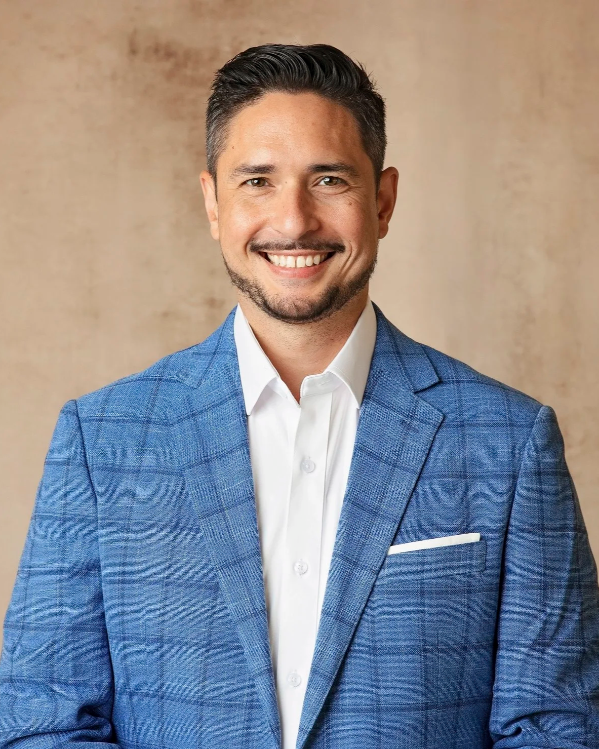 Paul smiling and wearing a white shirt and blue checkered blazer standing against a beige background.