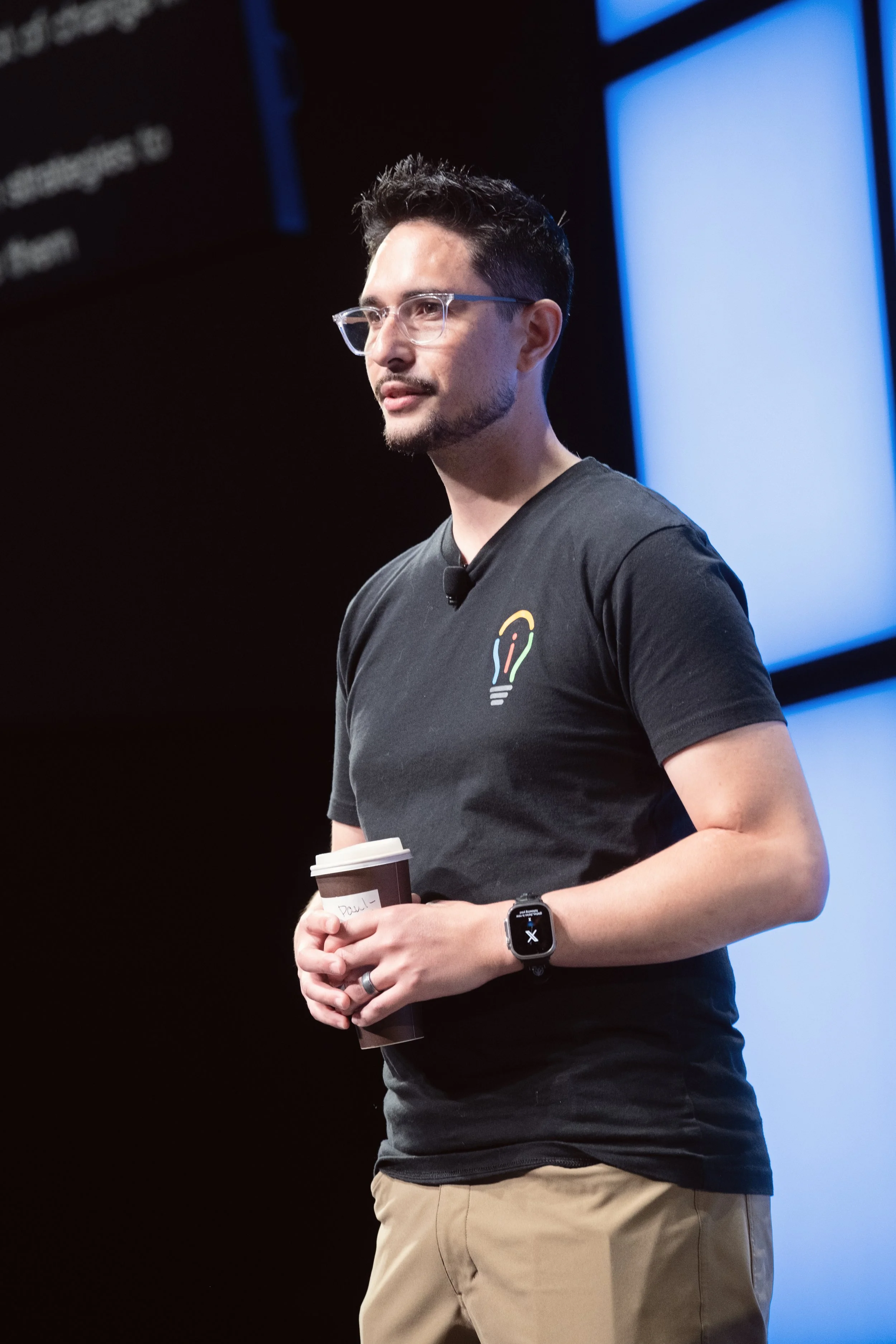 Paul with glasses holding a coffee cup, wearing a black t-shirt with a lightbulb logo, standing in front of a dark background with blue illuminated panels.
