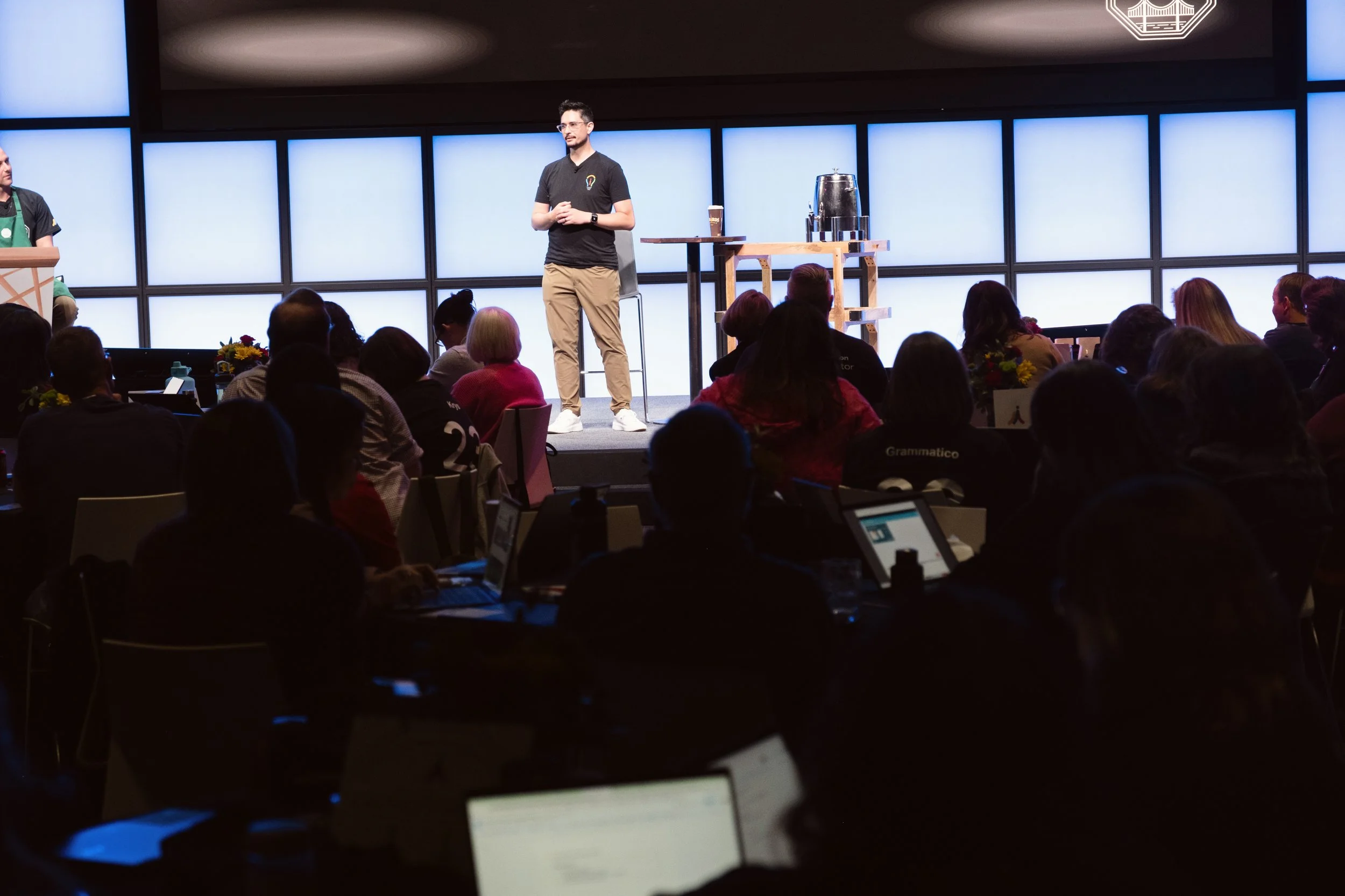 Paul is standing on a stage giving a presentation to an audience seated at tables. There are coffee beverages and a table with a metal container on stage. The background consists of large panels with illuminated sections.