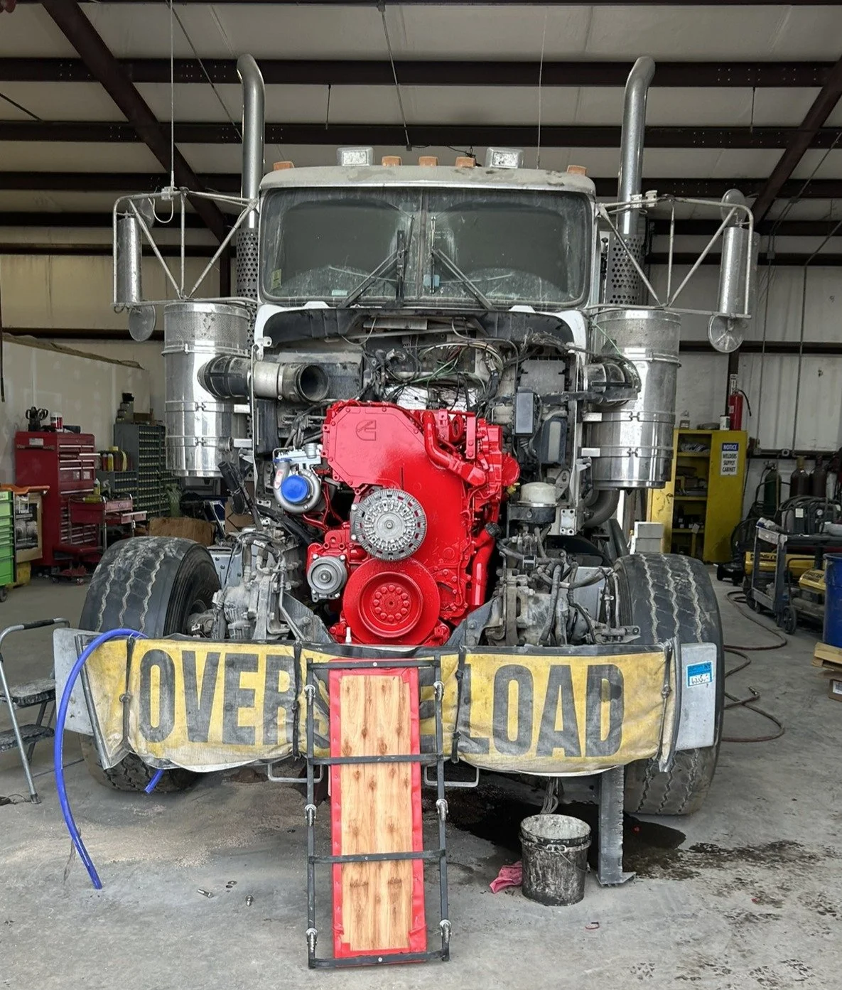Front view of a large truck with its engine exposed, showing a red engine block and various mechanical parts. The truck is in a workshop with tools and equipment in the background. A yellow caution sign saying 'Overload' is attached to the front bumper.