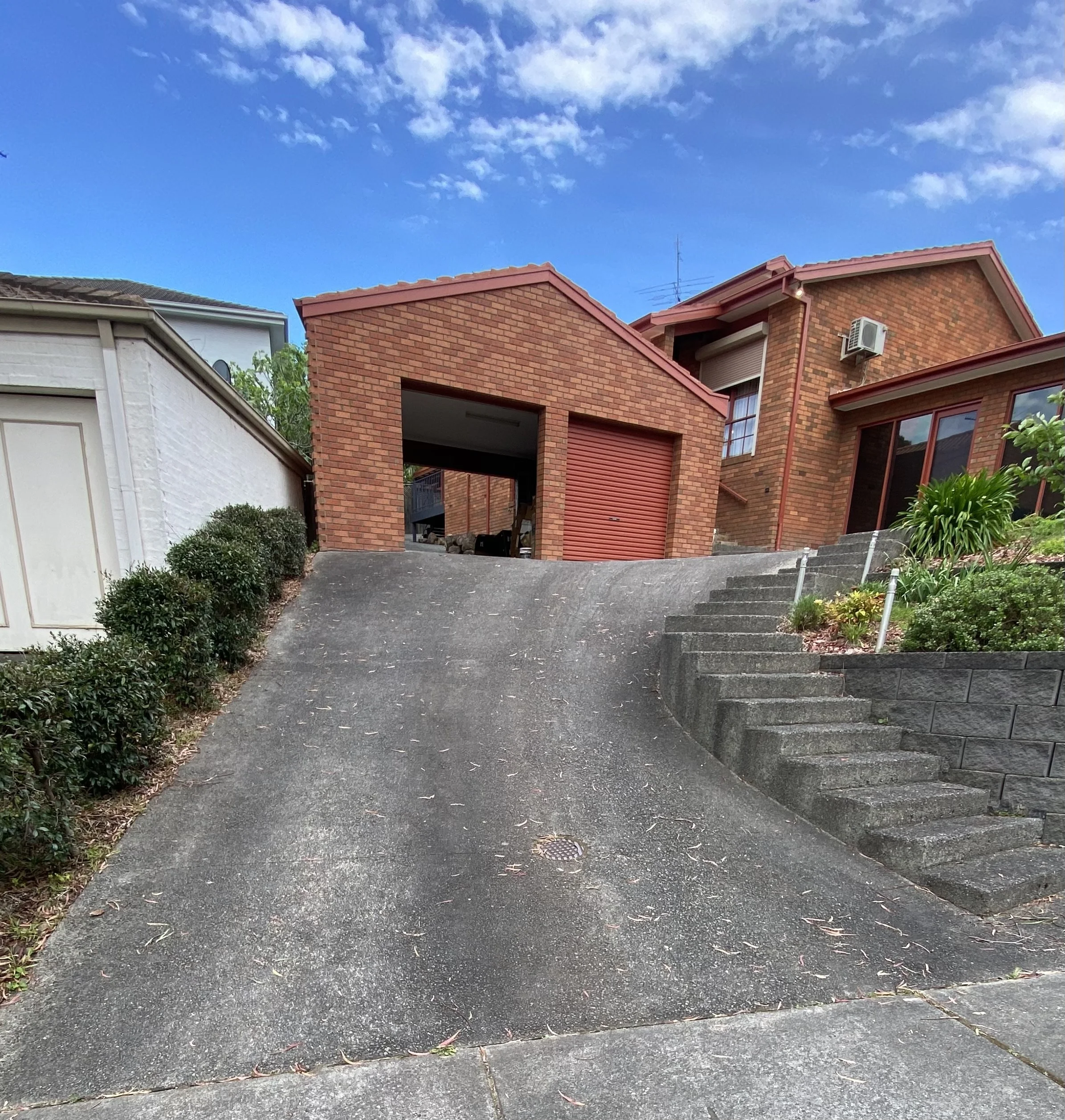 Residential house with brick and stucco exterior, driveway with stairs, surrounded by bushes and trees under a blue sky with some clouds.