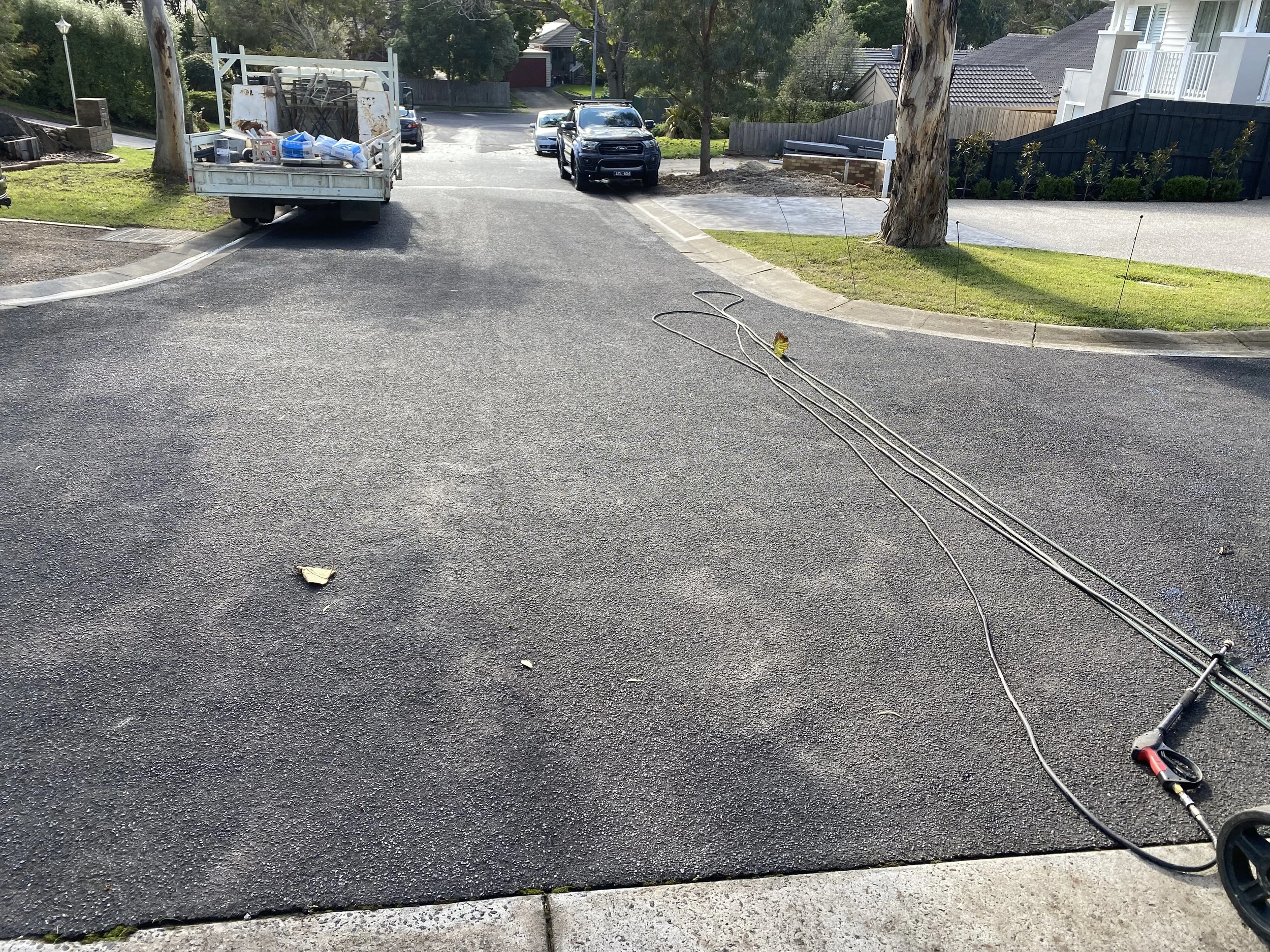 Residential driveway with construction tools and materials, including a power drill and extension cords, near a small patch of grass and two trees, with parked cars and a house in the background.