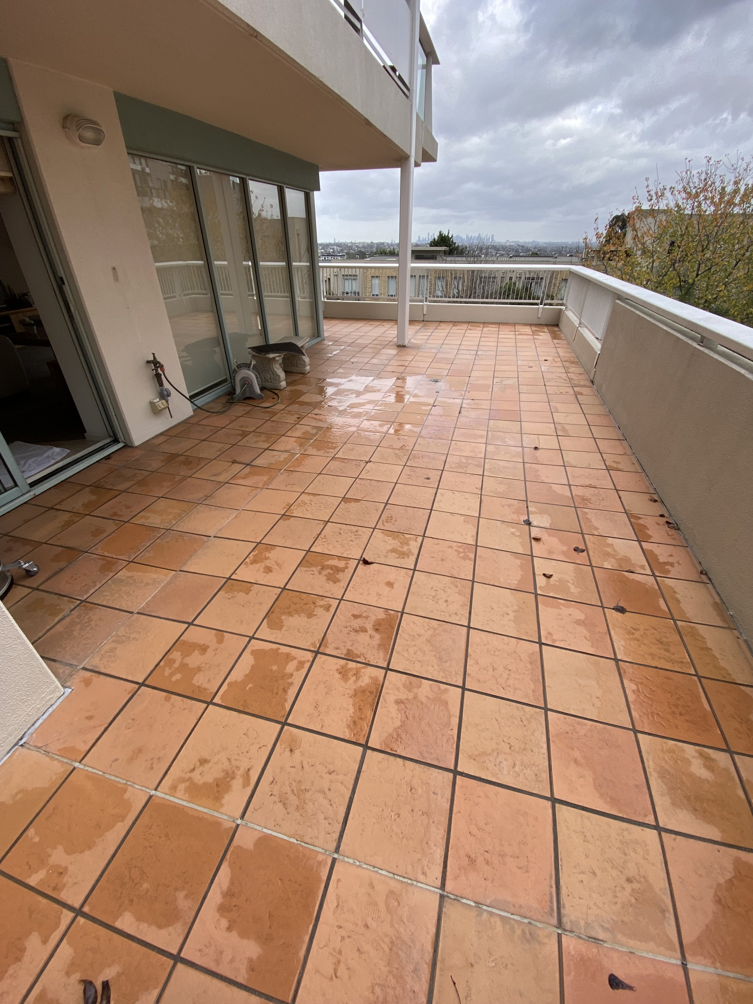 A large outdoor balcony with wet, orange-tiled flooring and a white railing. Part of the balcony has sliding glass doors leading inside a building. There are two concrete blocks and a hose reel on the left side near the wall. The sky is overcast with gray clouds, and distant city skyline can be seen on the horizon.