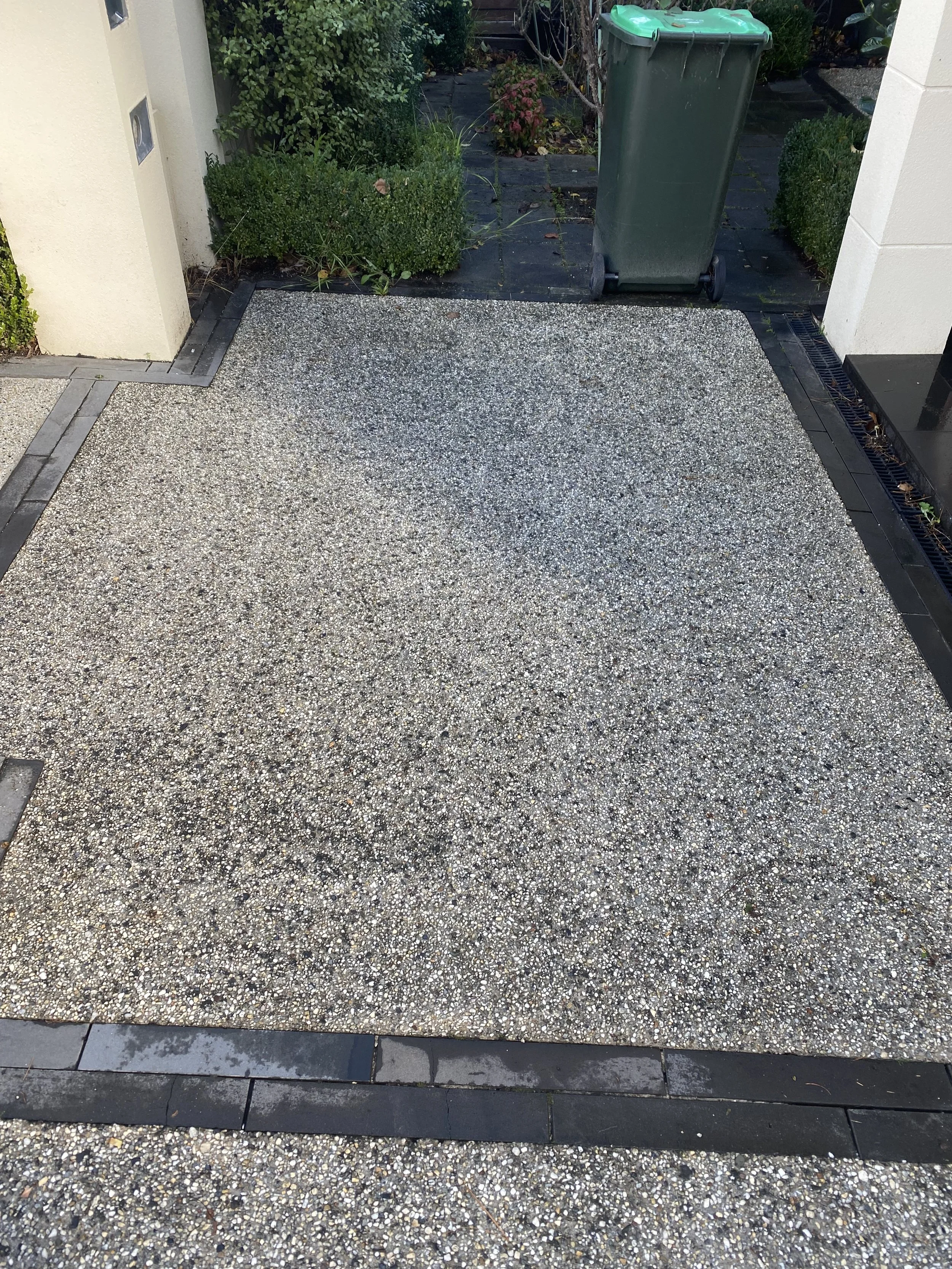 An outdoor area featuring a speckled marble-like tiled ground, bordered with black border stones. On the left side, part of a cream-colored structure with small square vents. In the background, there are green bushes and plants, and a gray pathway. A green trash bin with wheels is positioned on the pathway to the right.