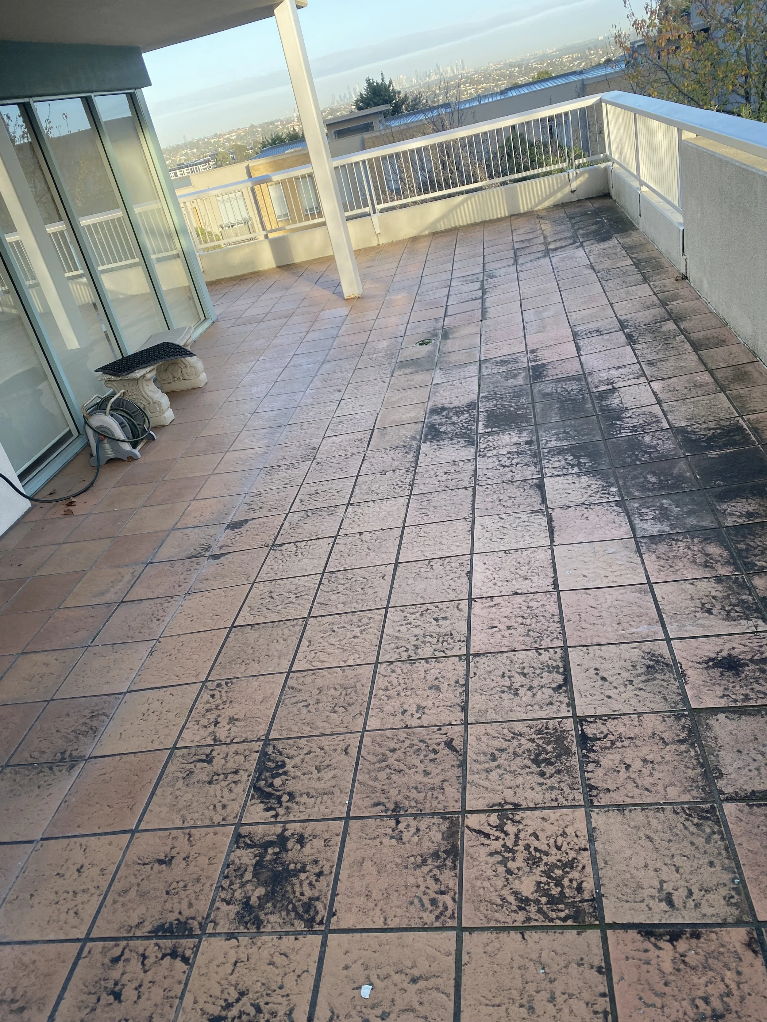 A spacious balcony or terrace with tiled flooring, located in a high-rise building with city views in the background. There are sliding glass doors on the left side, and the balcony has a white railing around it. Some cleaning tools are visible on the left side, and there are shadows cast on the floor from the railing and the building's structure.