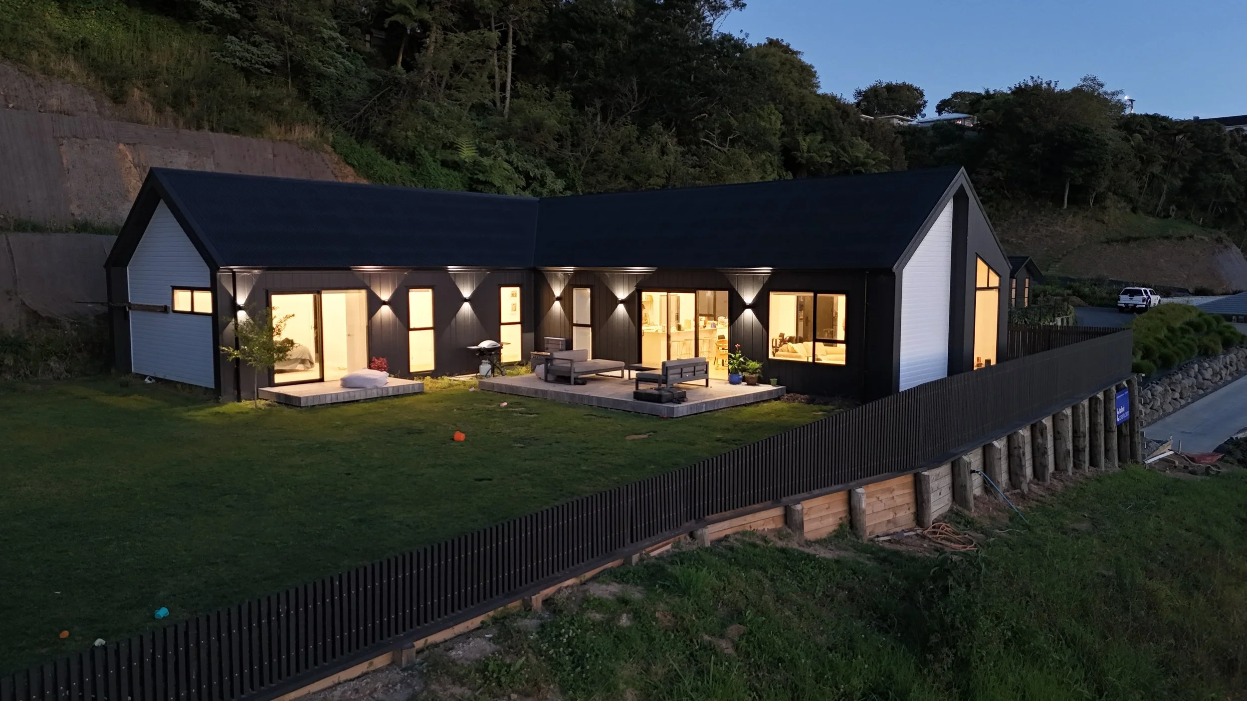 A modern house with large illuminated windows and a cozy outdoor patio area, set against a hillside at dusk.