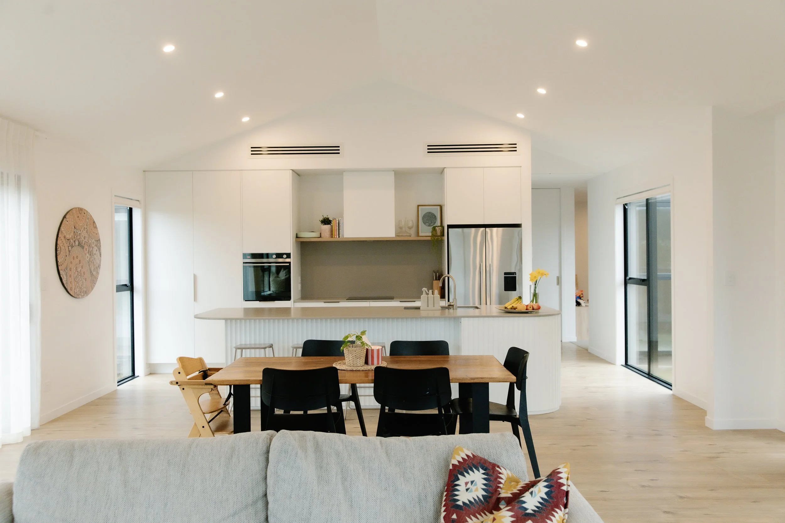Modern open-plan kitchen and dining area with white cabinets, stainless steel refrigerator, and a wooden dining table with black chairs. The space has large windows and sliding glass doors, light wood flooring, and minimal decorative accents.