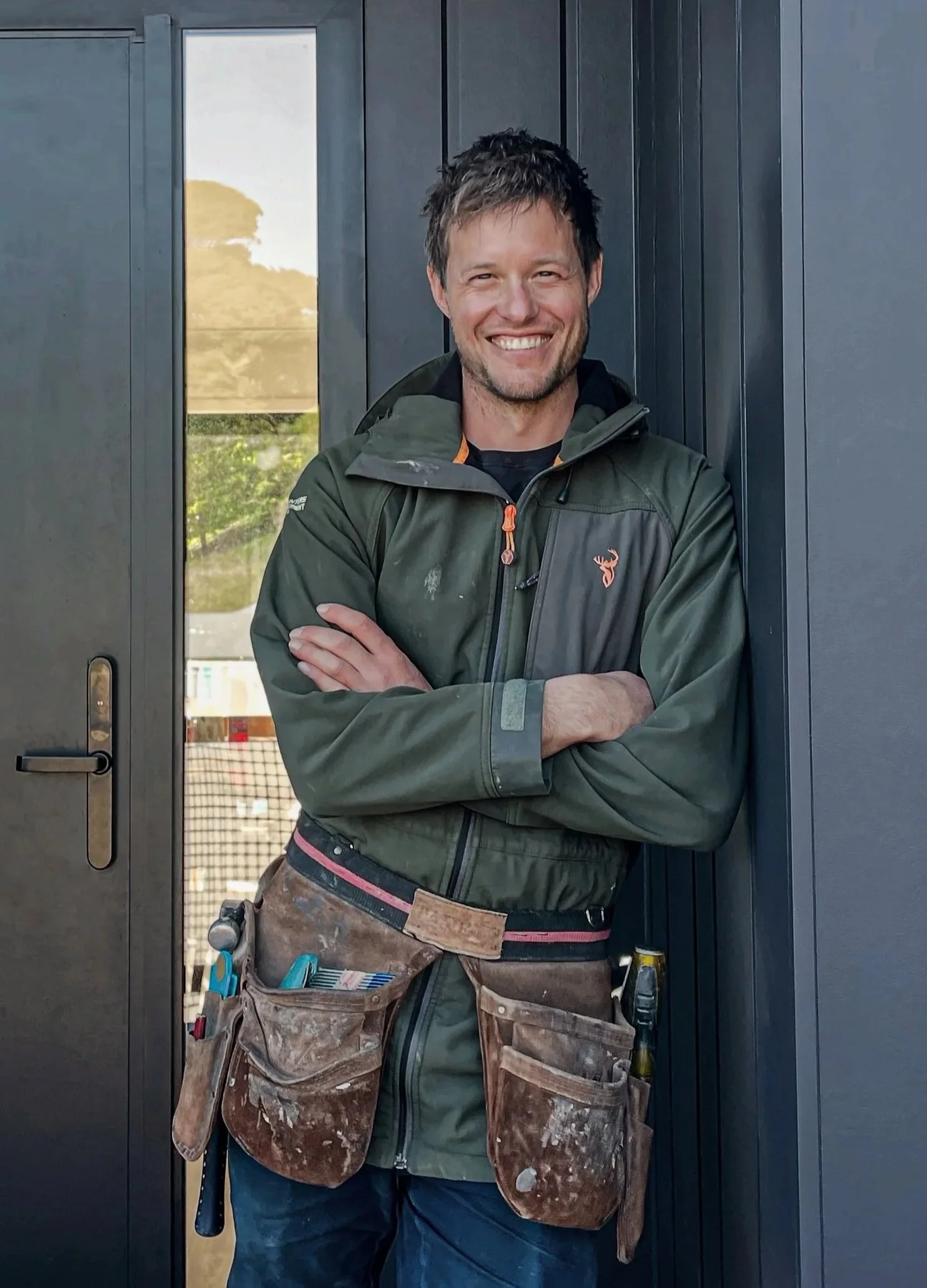 Man smiling, standing against dark gray door, wearing a green jacket with a logo, tool belt with various tools, and camouflage pants.