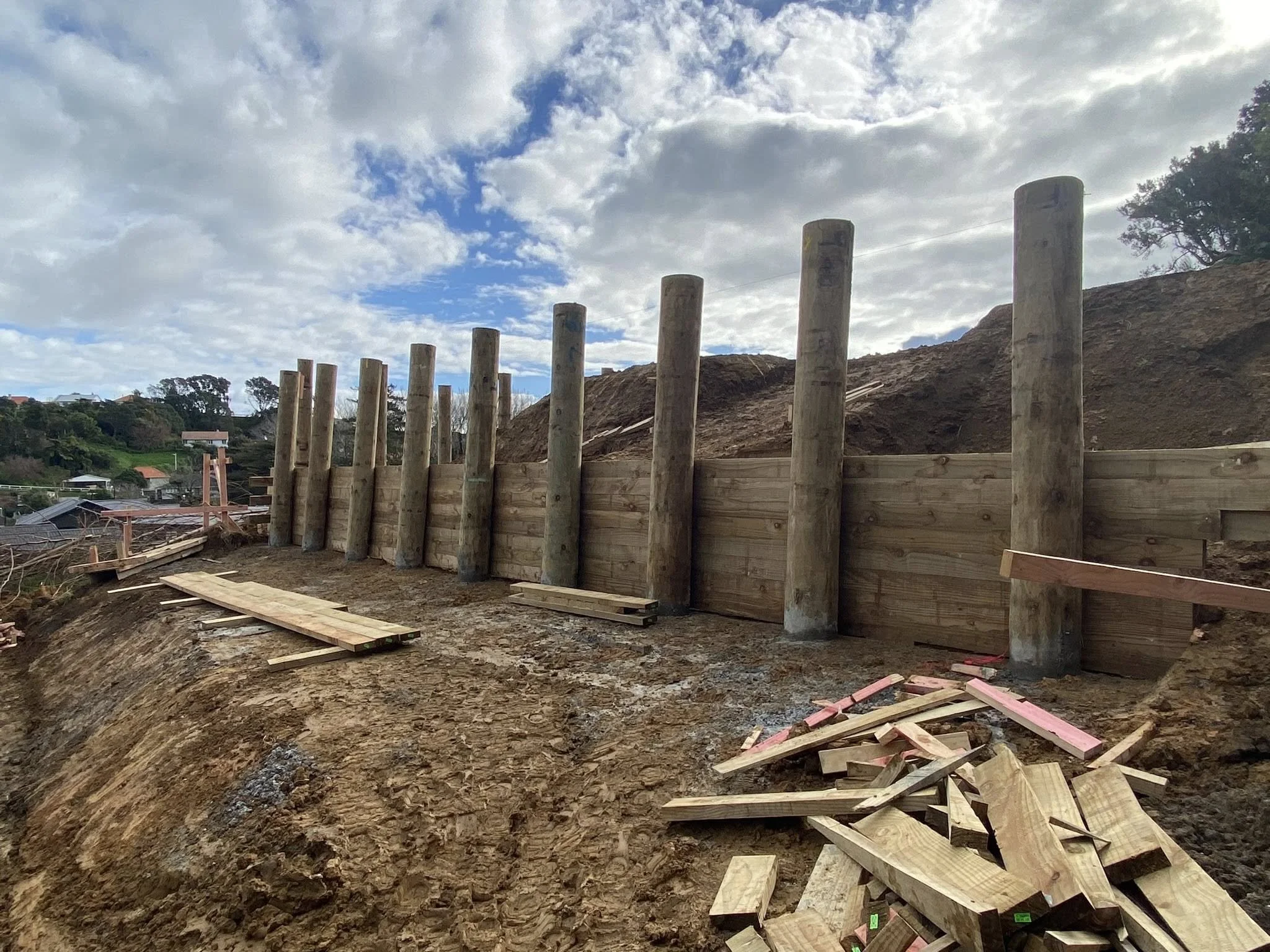 Construction site with wooden posts and a wooden fence, against cloudy sky and distant trees and hills.