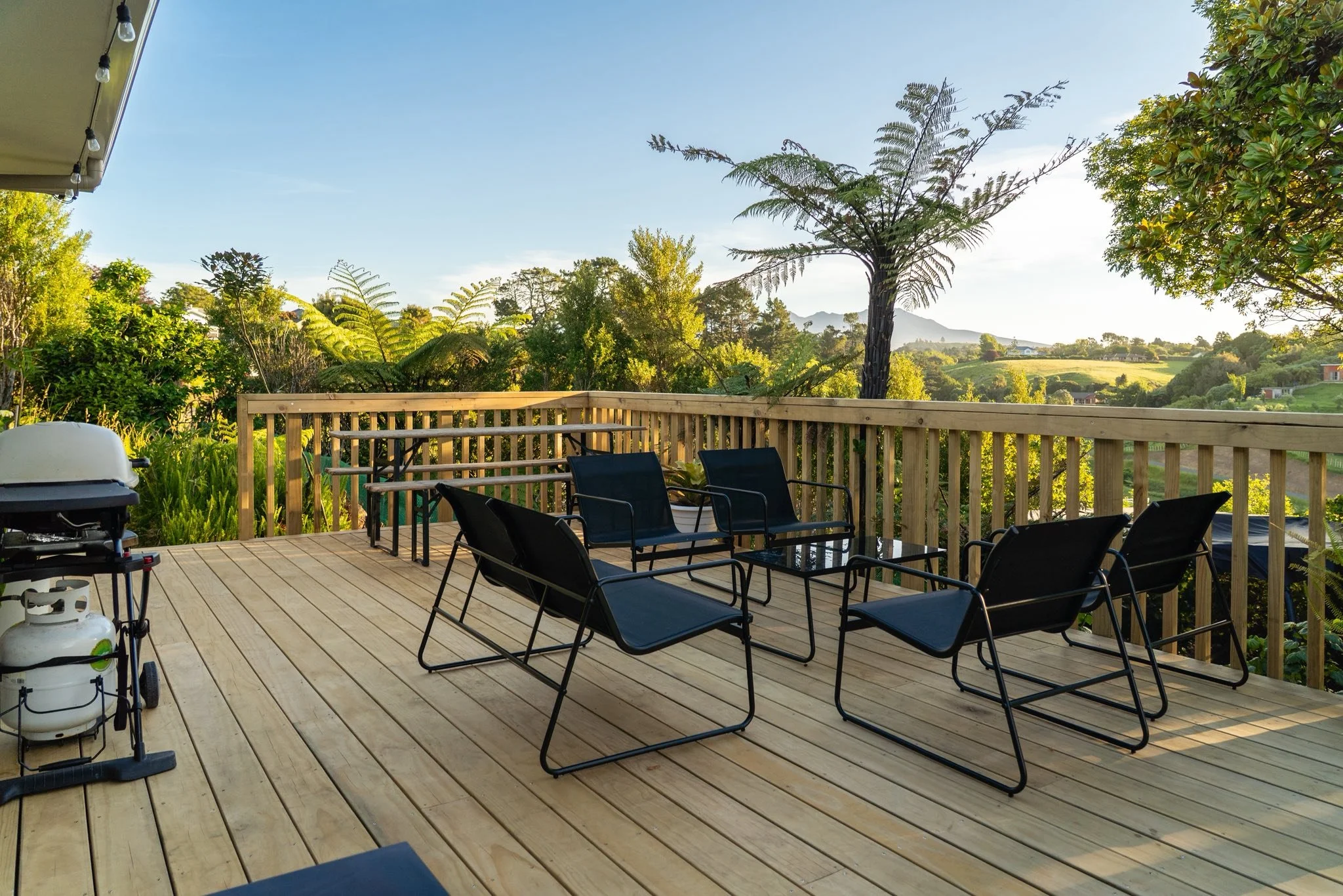 A wooden deck with black outdoor chairs, a grill, and propane tanks in the corner. A wooden railing surrounds the deck, overlooking a lush green landscape with trees and hills under a clear blue sky.