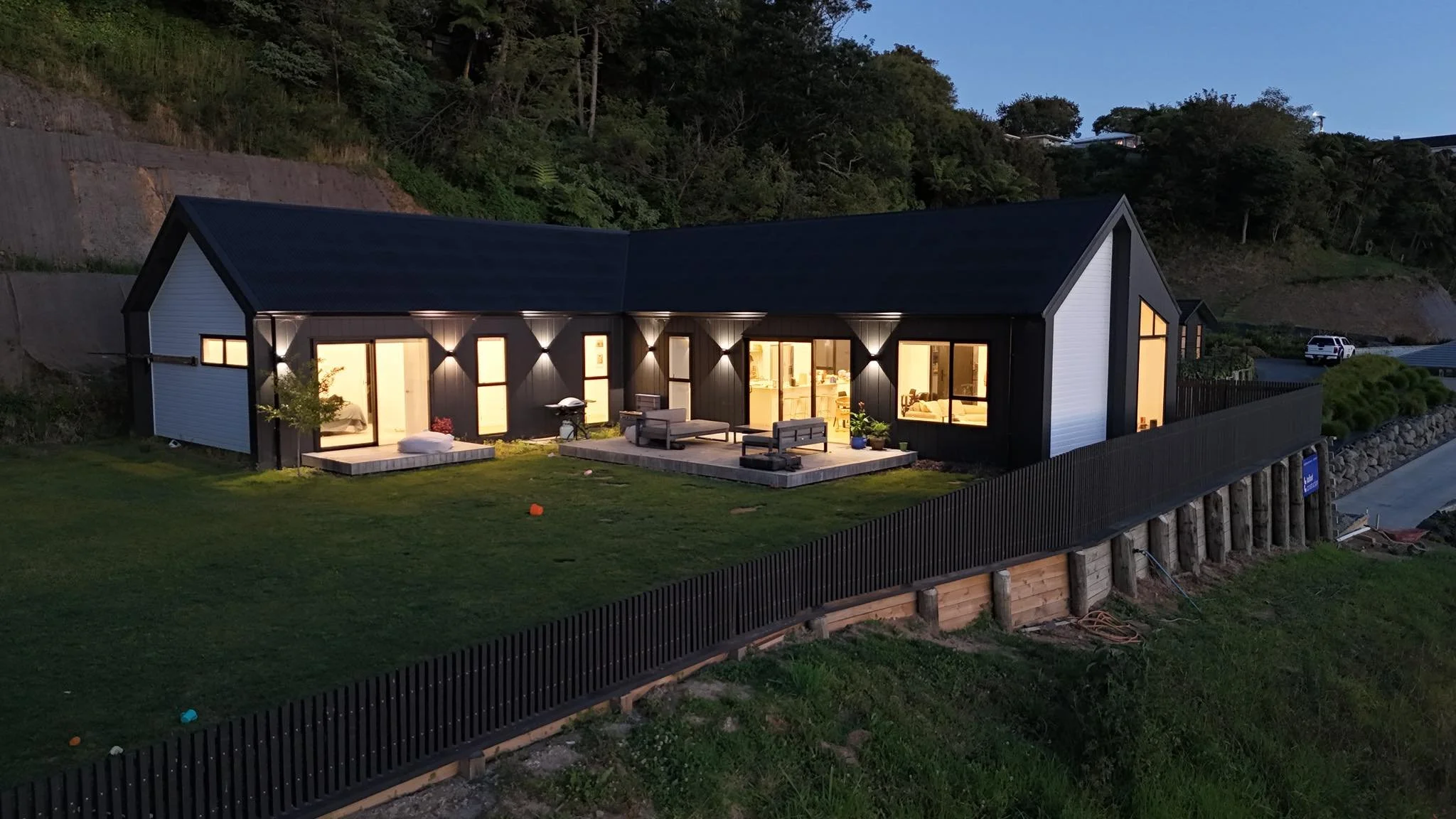 A modern house at dusk with warm interior lighting visible through large windows and sliding glass doors. There is a small patio with outdoor furniture, a barbecue grill, and a grassy yard. The house is built on a hillside with retaining walls and is surrounded by forested terrain.