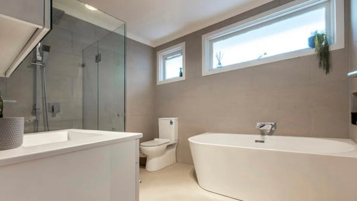 A modern bathroom featuring a bathtub, toilet, glass shower enclosure, and small window with plants on the sill.