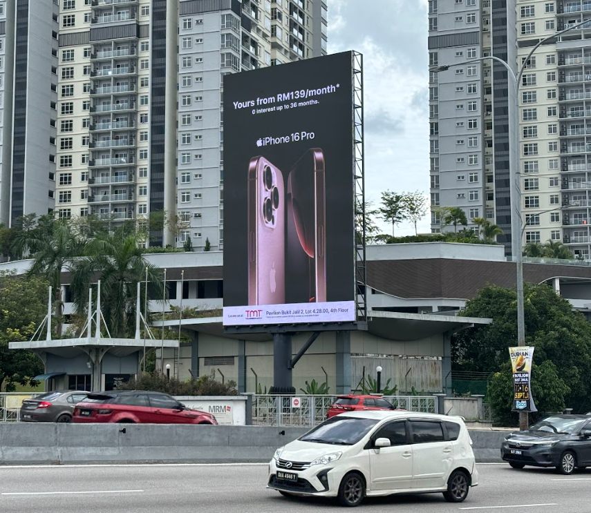 Large outdoor billboard advertising the iPhone 16 Pro, showing a pink phone with text about pricing and interest rate in a city with high-rise apartment buildings and cars on the road.