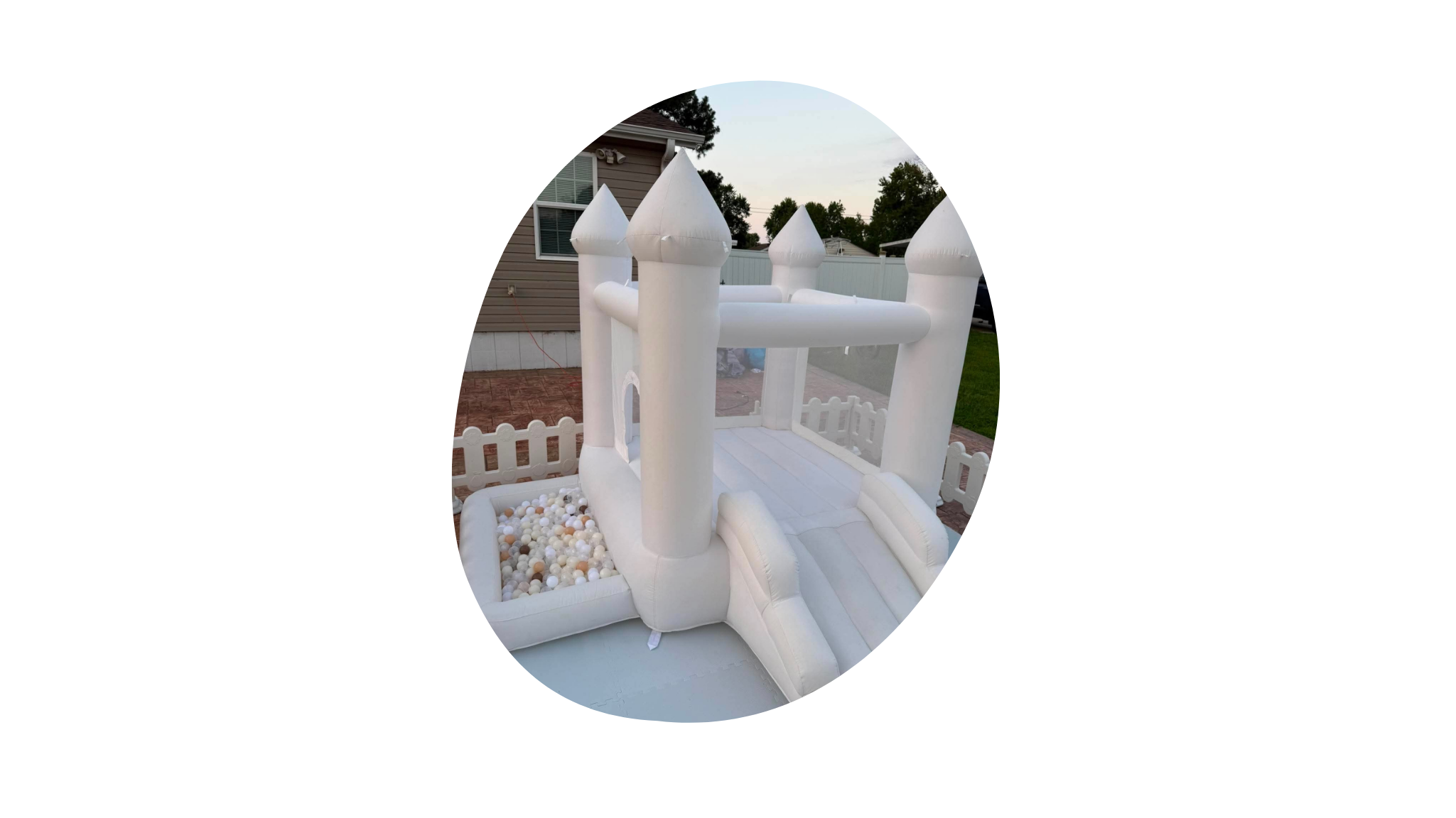 An inflatable white castle bounce house with a slide, located outdoors on a residential backyard patio, with a house and trees in the background.