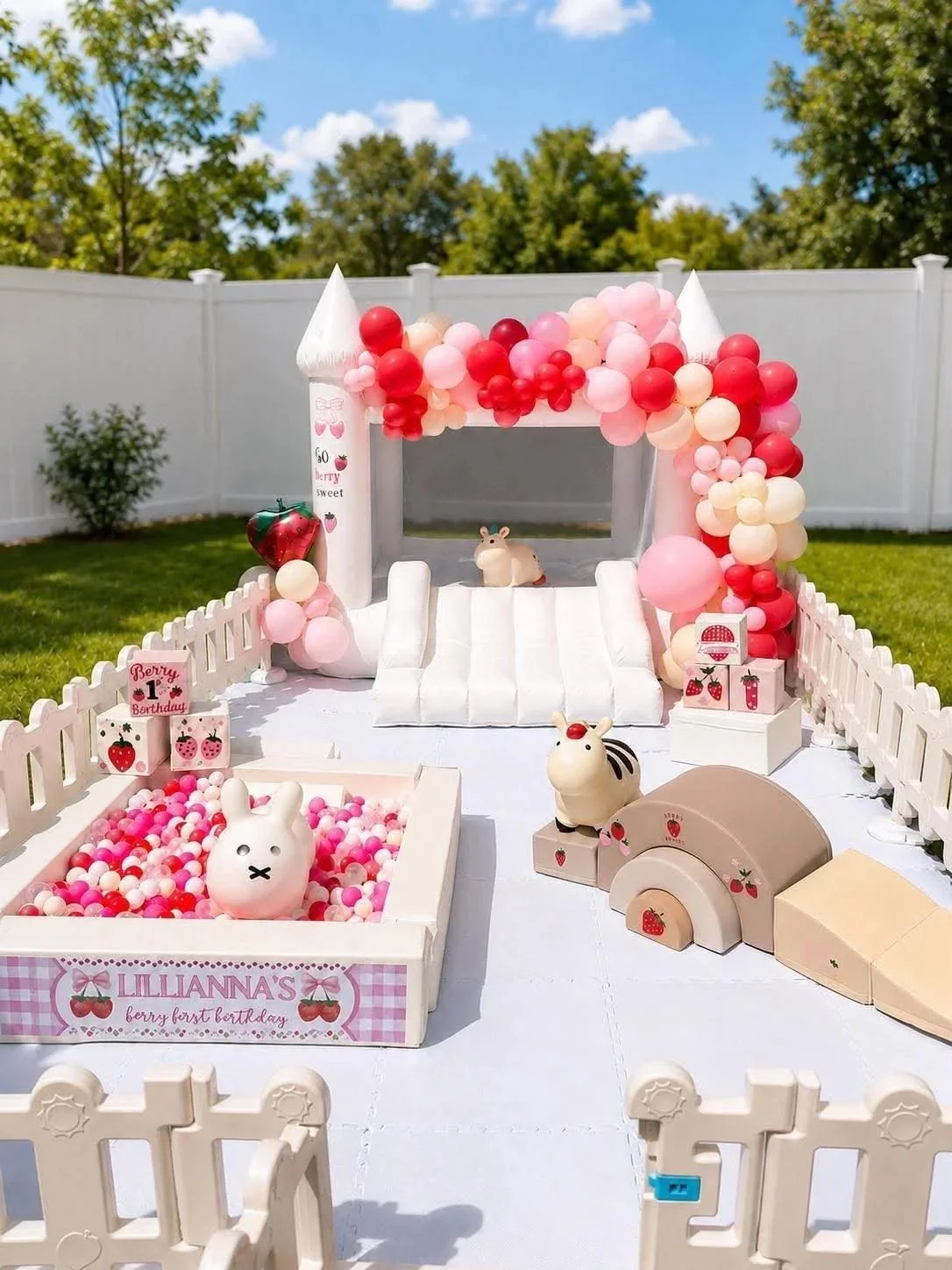 Decorative outdoor setup for a toddler's first birthday party with a bunny theme, featuring a white fence, pink and red balloons, small animal figurines, a ball pit filled with pink and white balls, and birthday signs.