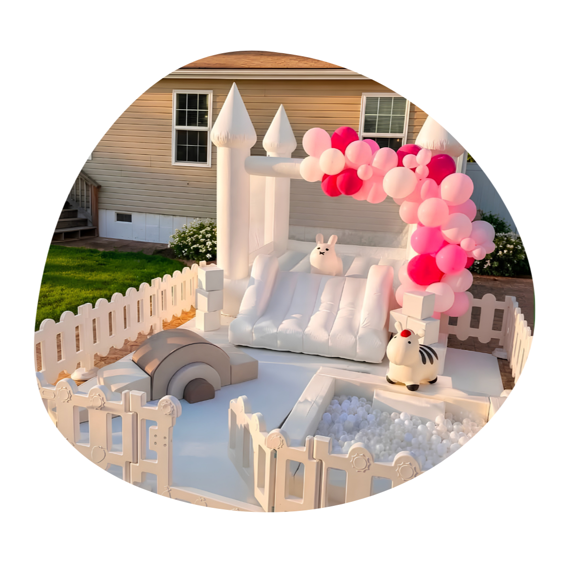 A white inflatable bounce house with a castle theme decorated with pink and white balloons, featuring plush bunny and zebra toys, and surrounded by a white picket fence, set outdoors on a lawn against a beige house.