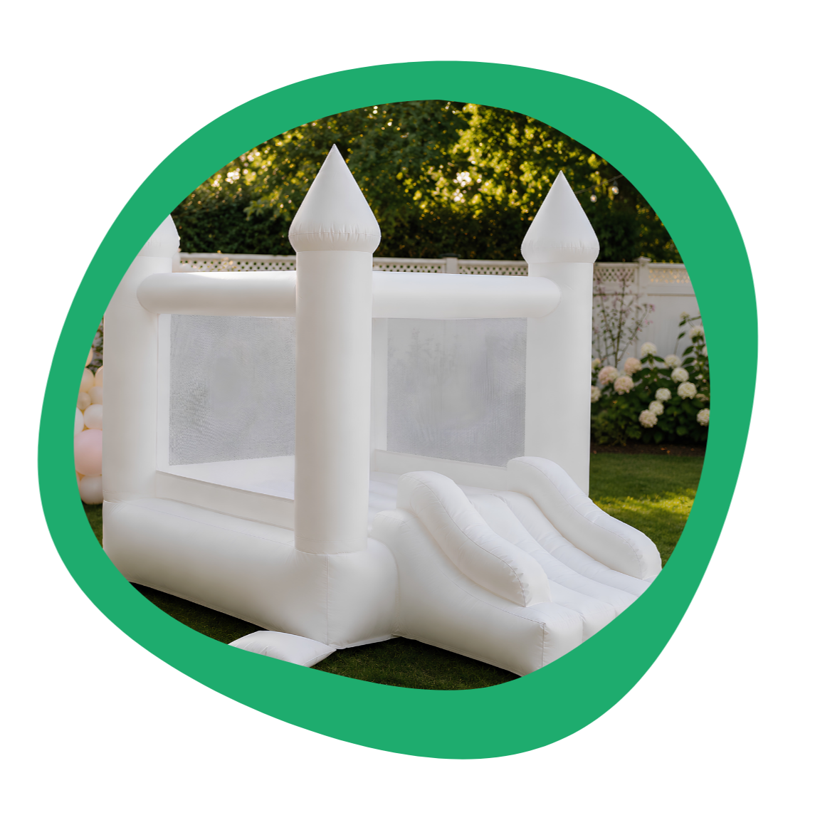 White inflatable bounce house castle with slide, set outdoors on grass with trees and flowers in the background.