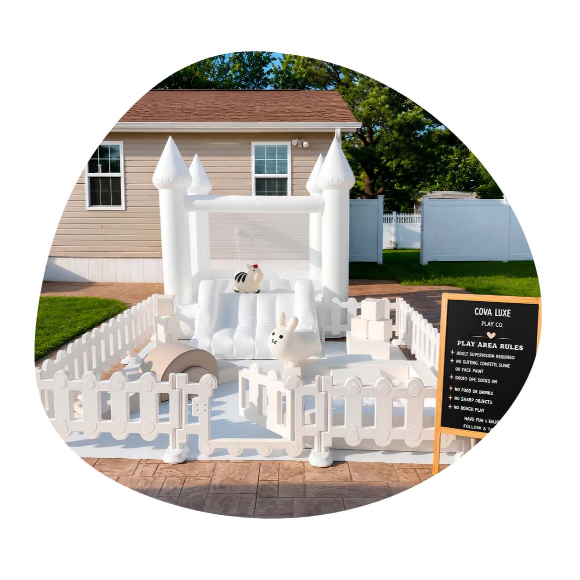 A white inflatable bounce house with castle towers, set up in a backyard with grass and fencing. It has plush animal toys inside and a blackboard with play area rules.