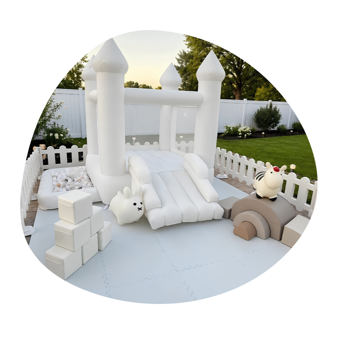 Inflatable white castle with slide, fenced yard, outdoor toys including a rabbit-shaped figure and a zebra-shaped figue, grass, trees, and bushes.