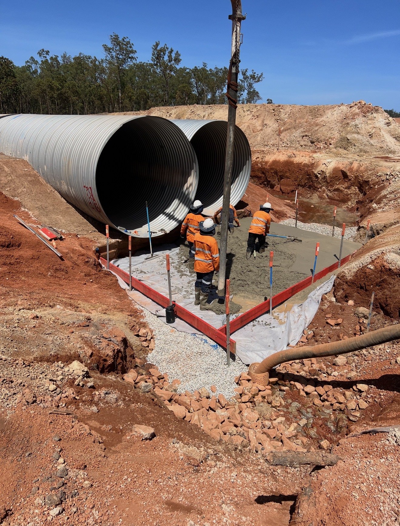 Construction workers in orange safety vests and hard hats working on a large pipeline installation at a construction site with orange dirt and gravel.