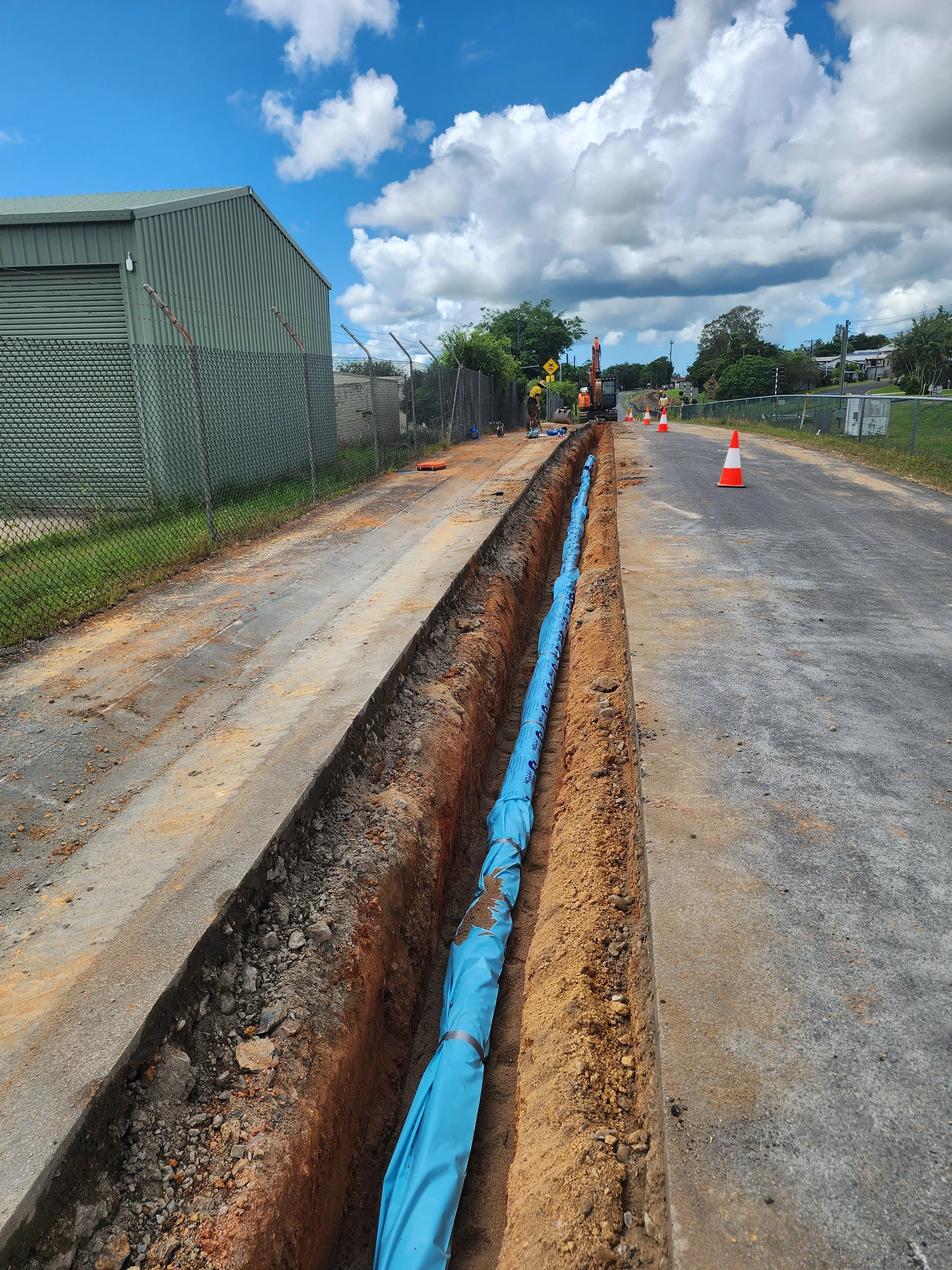 Road construction scene with workers installing underground utility pipes, traffic cones, and machinery under a partly cloudy sky.