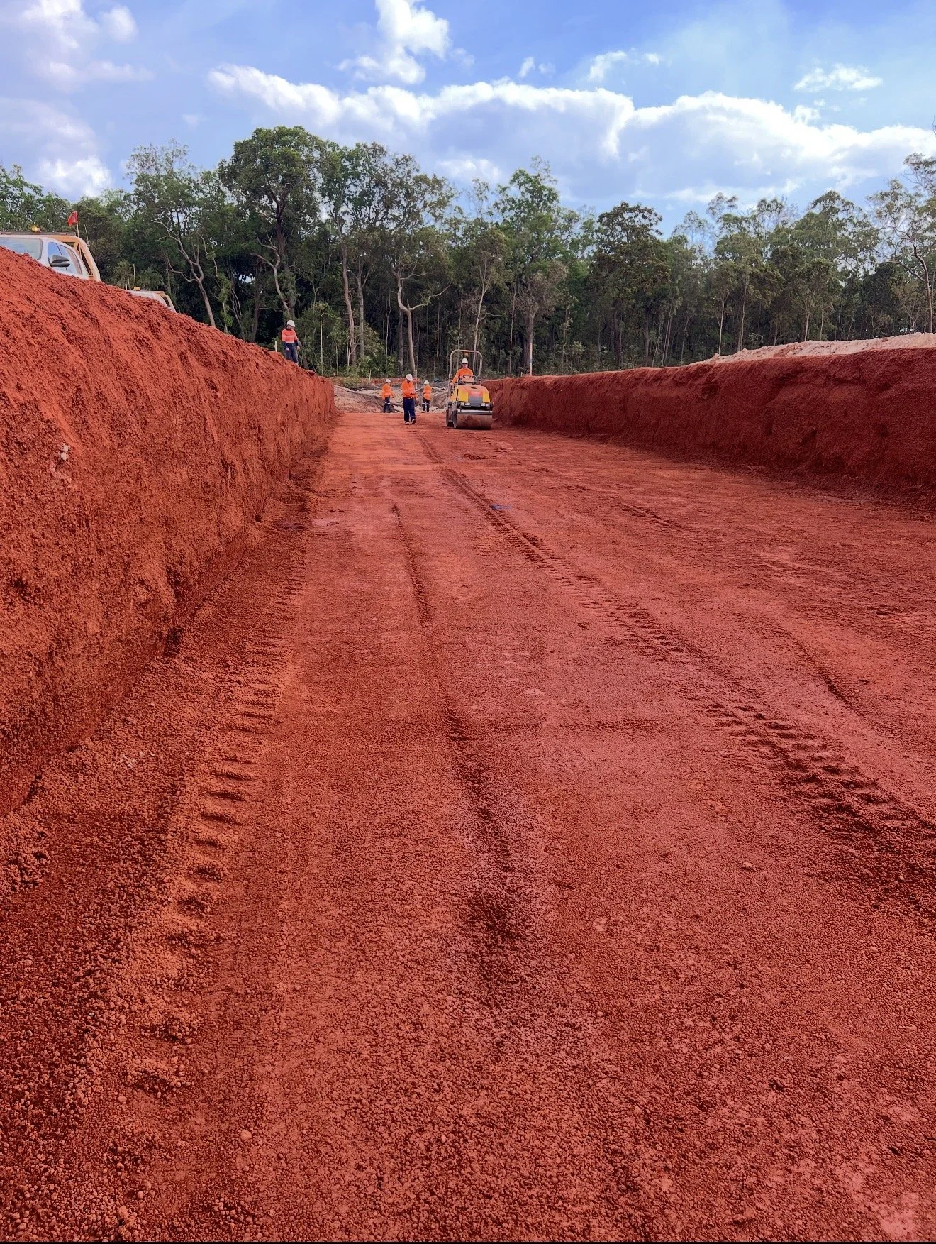 Construction workers working on a red dirt road with large dirt walls on either side, surrounded by a forested area and a partly cloudy sky.