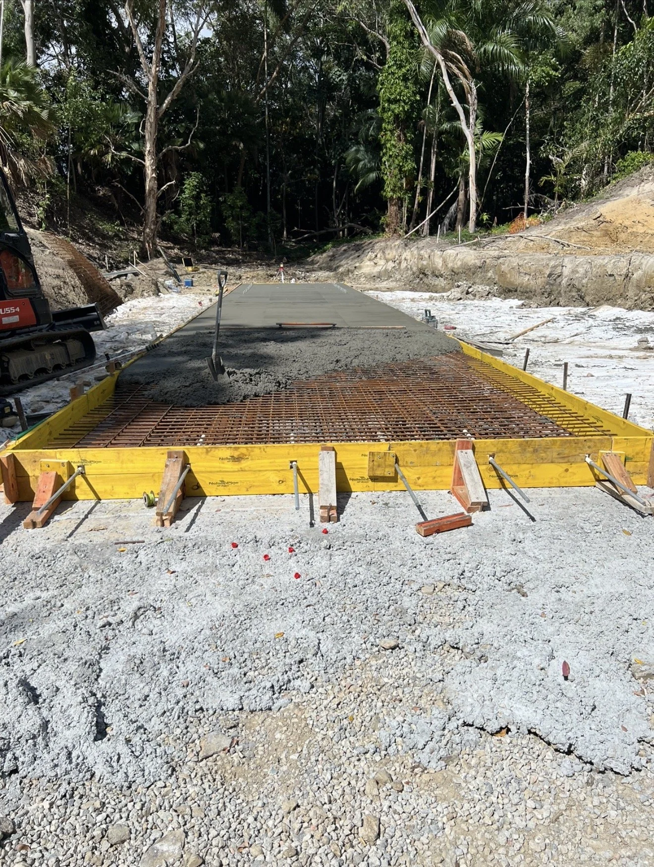 Construction site with a concrete slab being poured on a gravel foundation, surrounded by lush green trees.