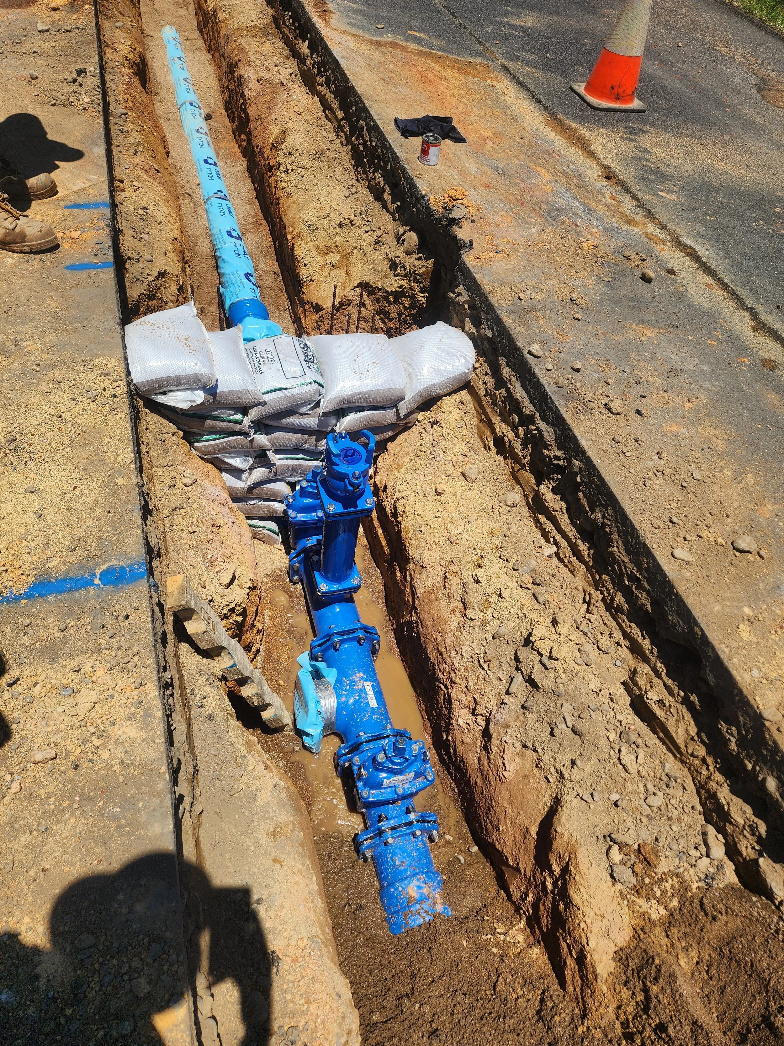 Construction site where a blue pipe is being installed in a trench with construction materials, a traffic cone, and tools nearby.