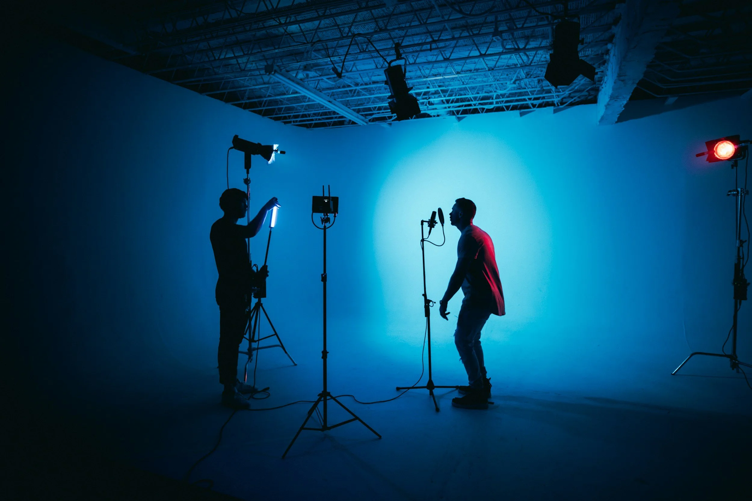 Two people in a photography studio setting with blue ambient lighting. One person is adjusting equipment while the other is standing near a microphone, with studio lights and cameras setup around them.