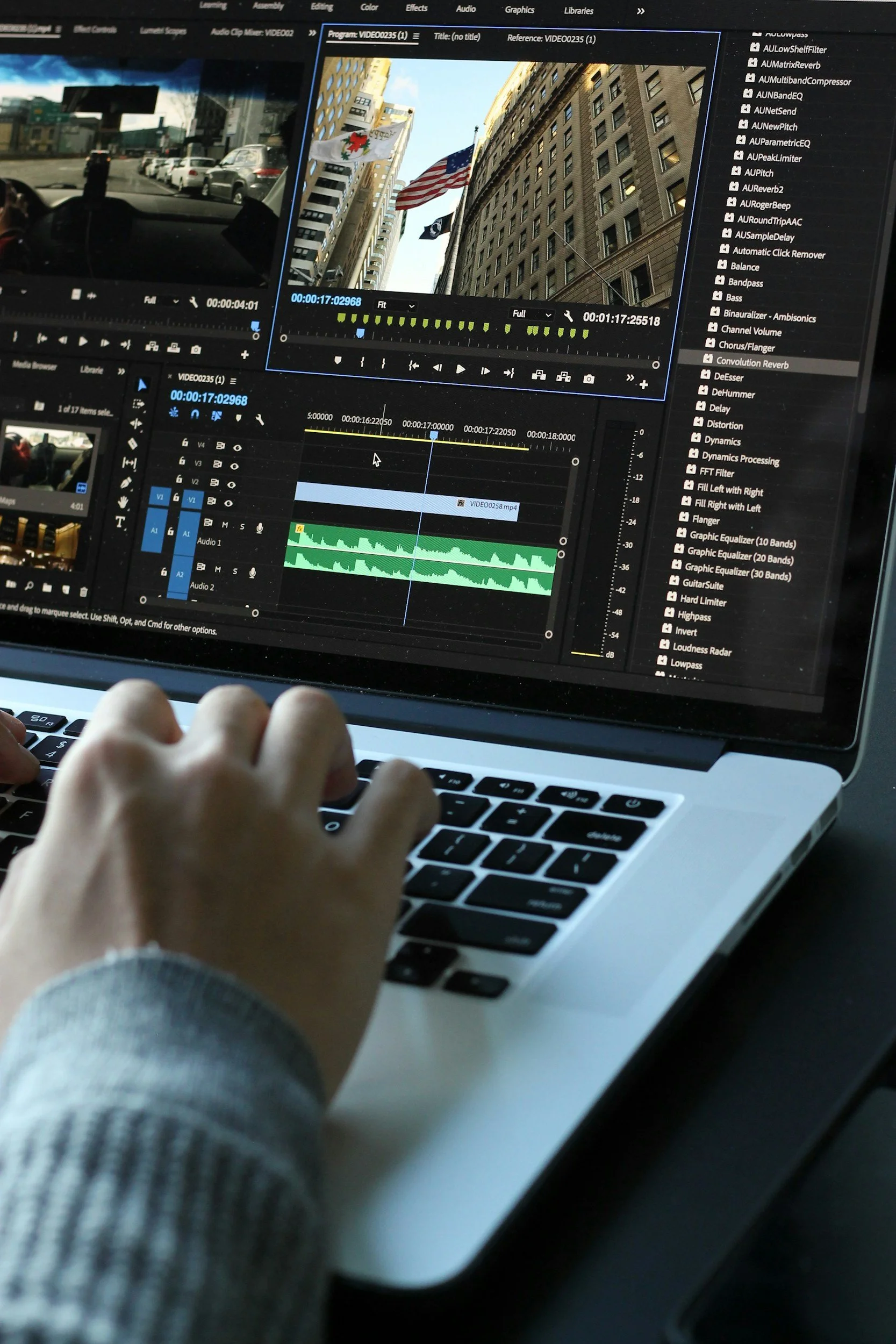A person working on a video editing software on a laptop, with architecture and flags of the United States and other flags on a building in the city visible through the screen reflection.