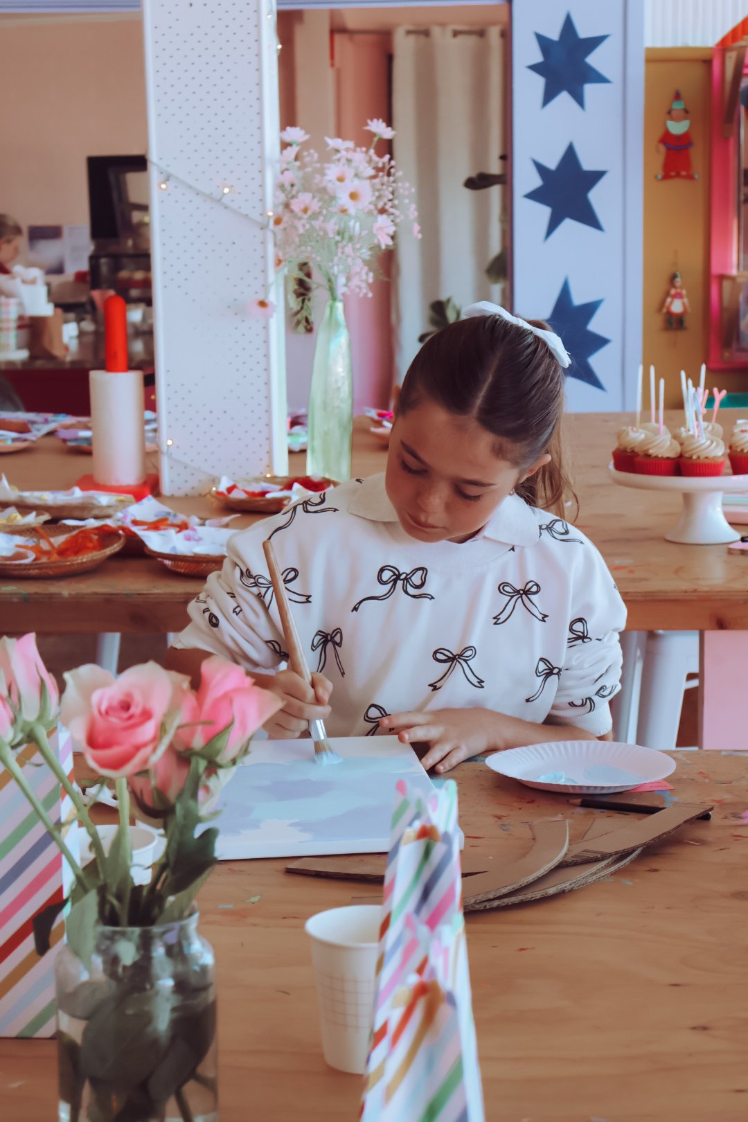 A young girl with a white bow in her hair, wearing a white blouse with black bow patterns, is painting at a wooden table decorated for a birthday party. The table has flowers in a jar, striped party hats, paper plates, and cupcakes with candles. In the background, there are colorful decorations, including a blue star banner and a display of pink flowers.