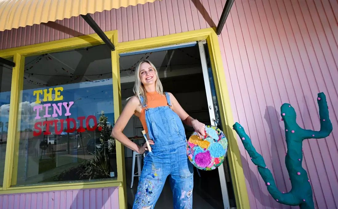 Young woman smiling inside an art studio holding a colorful painted canvas, standing in front of a glass door that reads "The Tiny Studio" with pink and blue walls and cactus decorations outside.
