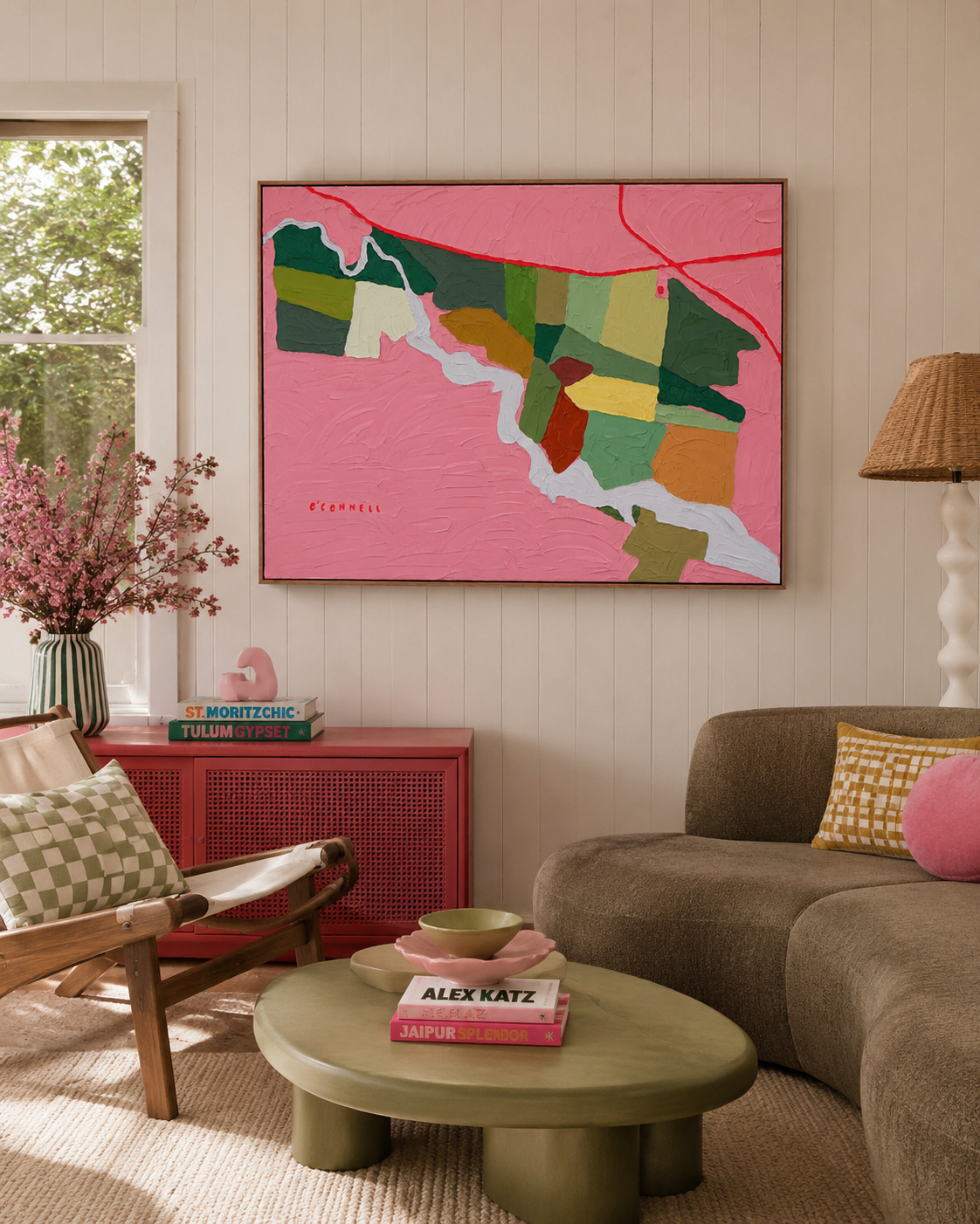 Interior of a living room with a beige sofa, a green coffee table with pink and green bowls, a patterned pillow, a red cabinet with books and decorative items, pink flowers in a vase, a large pink abstract painting on a light-colored wall, and a window with greenery outside.
