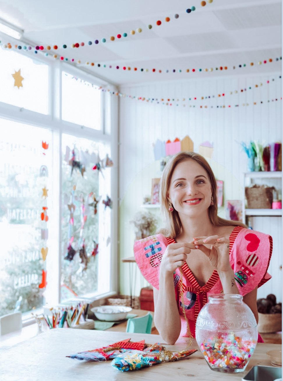 A woman smiling at a craft table decorated with colorful craft materials, in a room with window and hanging colorful decorations.