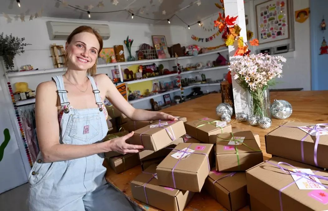 A woman smiling while holding a wrapped gift in a decorated room with a table full of more wrapped gifts, a vase of pink flowers, and holiday decorations.