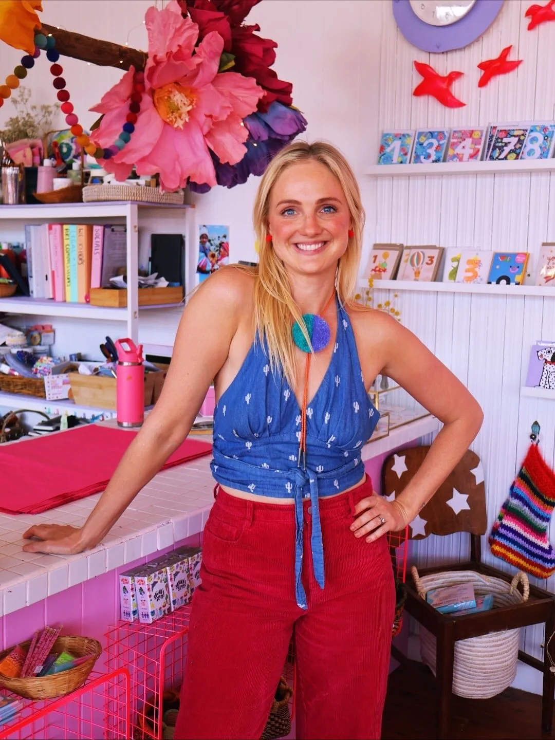 A woman smiling inside a colorful craft or gift shop decorated with flowers and numbers.