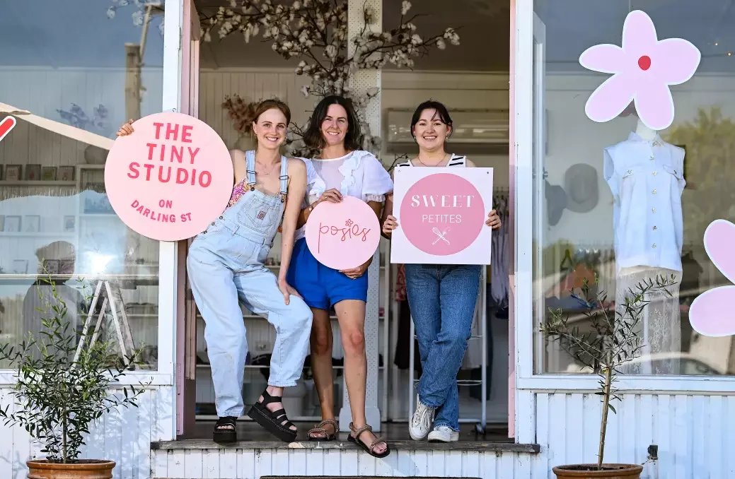 Three women standing at the entrance of a boutique store with pink signage and signs that read 'The Tiny Studio on Darling St,' 'Posies,' and 'Sweet Petites.' They are smiling and holding the signs, with floral decorations and mannequins visible inside.