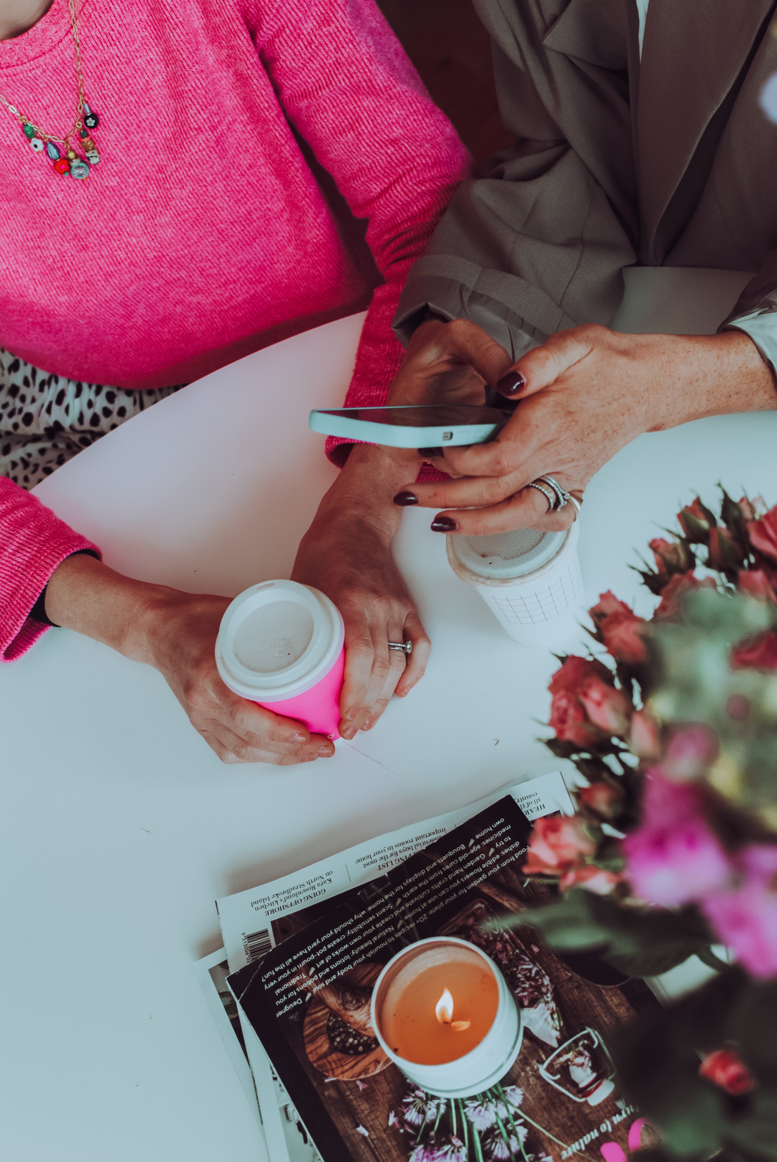 Two people are sitting at a white table, one wearing a pink sweater with a colorful necklace, and the other in a beige blazer. They are holding a smartphone together. On the table, there are two coffee cups, one pink and one white, a candle with a flame, and magazines or brochures. A bouquet of pink flowers is visible in the foreground.