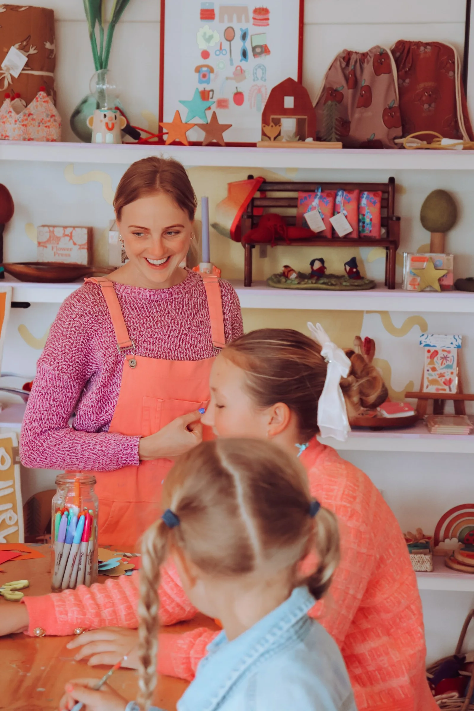 A woman with reddish-brown hair wearing a pink long-sleeve top and apron, smiling at children decorating crafts at a table in a classroom or craft room.