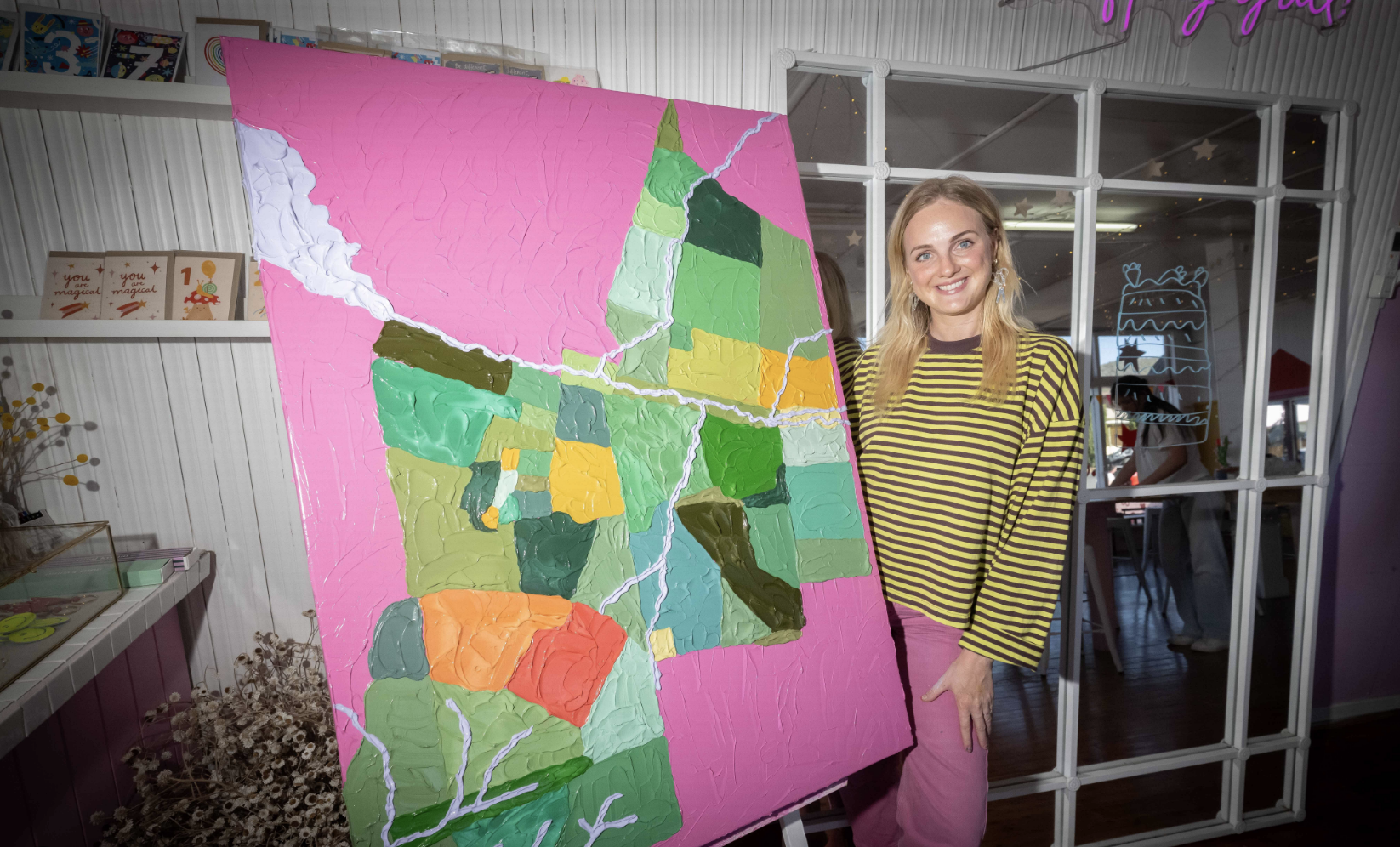 Woman posing next to a colorful abstract landscape painting on an easel inside a decorated room.