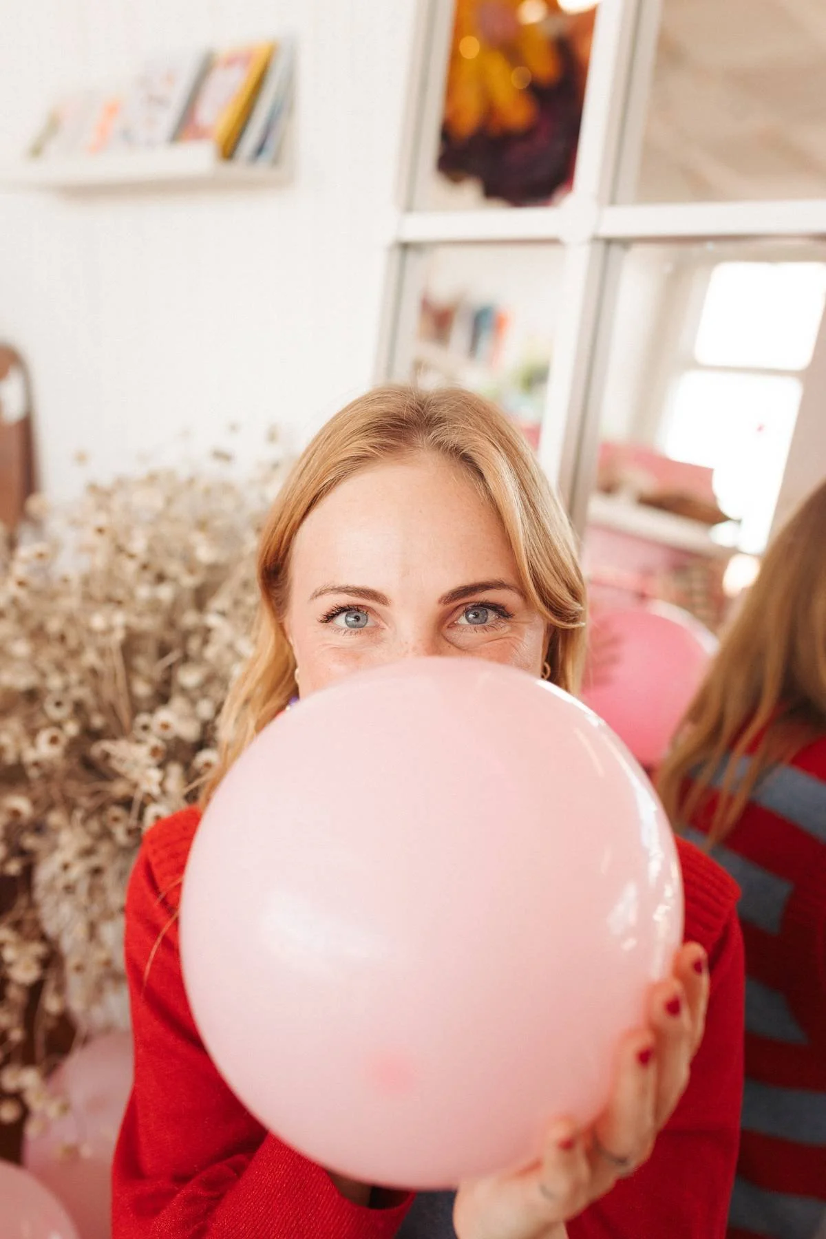 A woman with blonde hair and blue eyes holding a pink balloon, smiling and looking into the camera, wearing a red sweater, indoors with bookshelves and another person partially visible in the background.